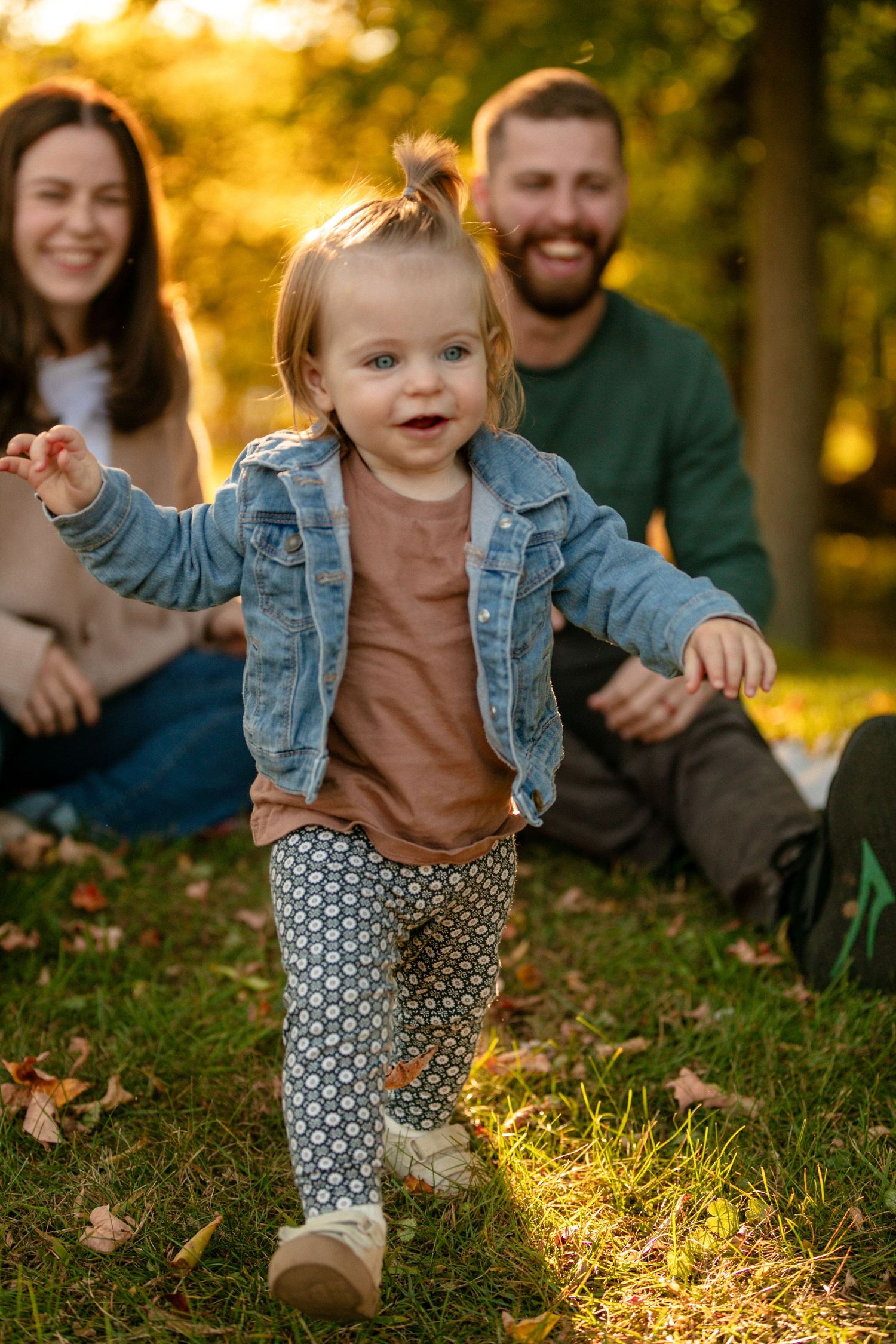 A toddler in denim jacket and patterned pants walks joyfully through fallen leaves at sunset.