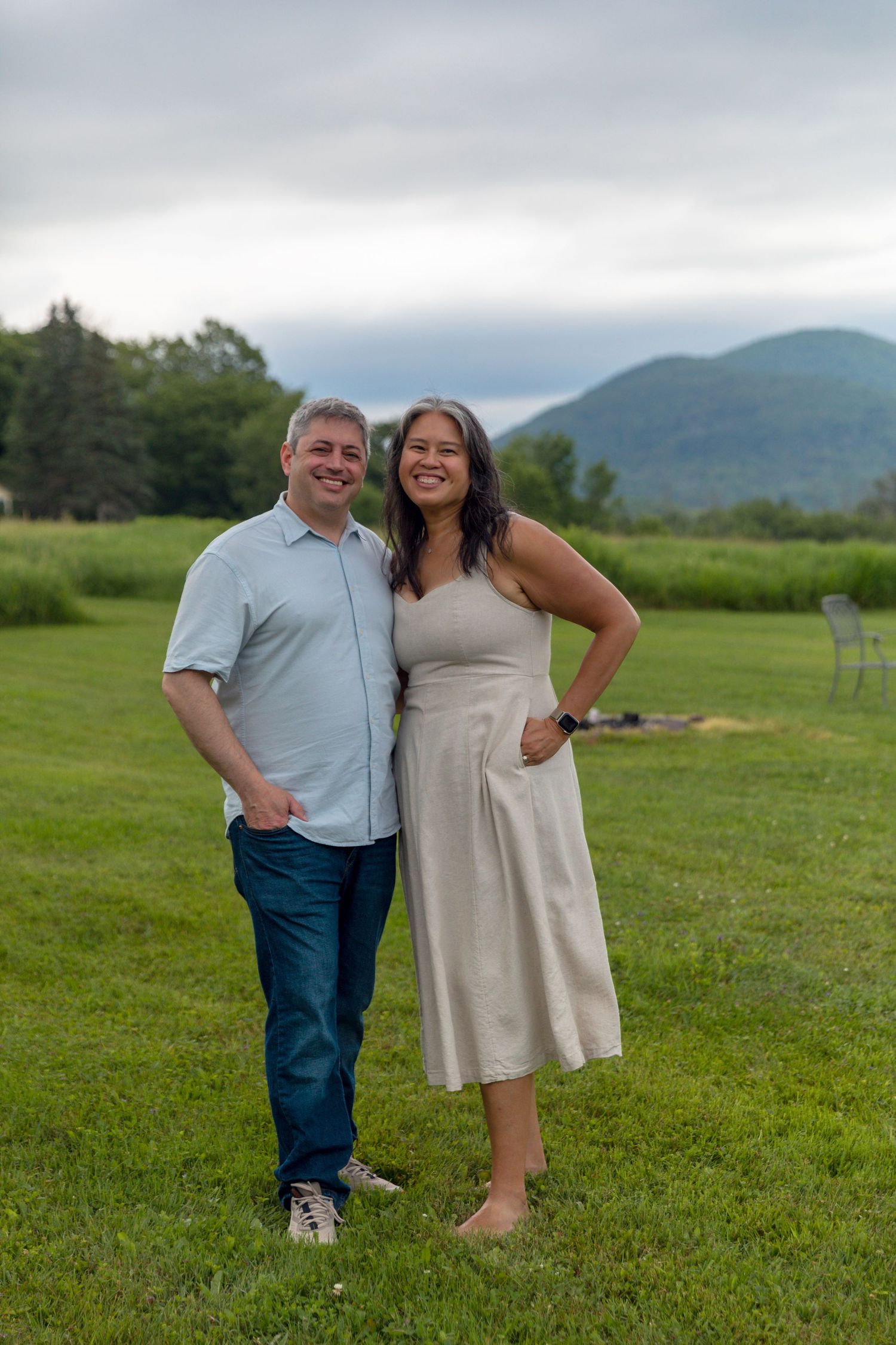 A couple poses together on a grassy field with mountains and cloudy skies in the background.