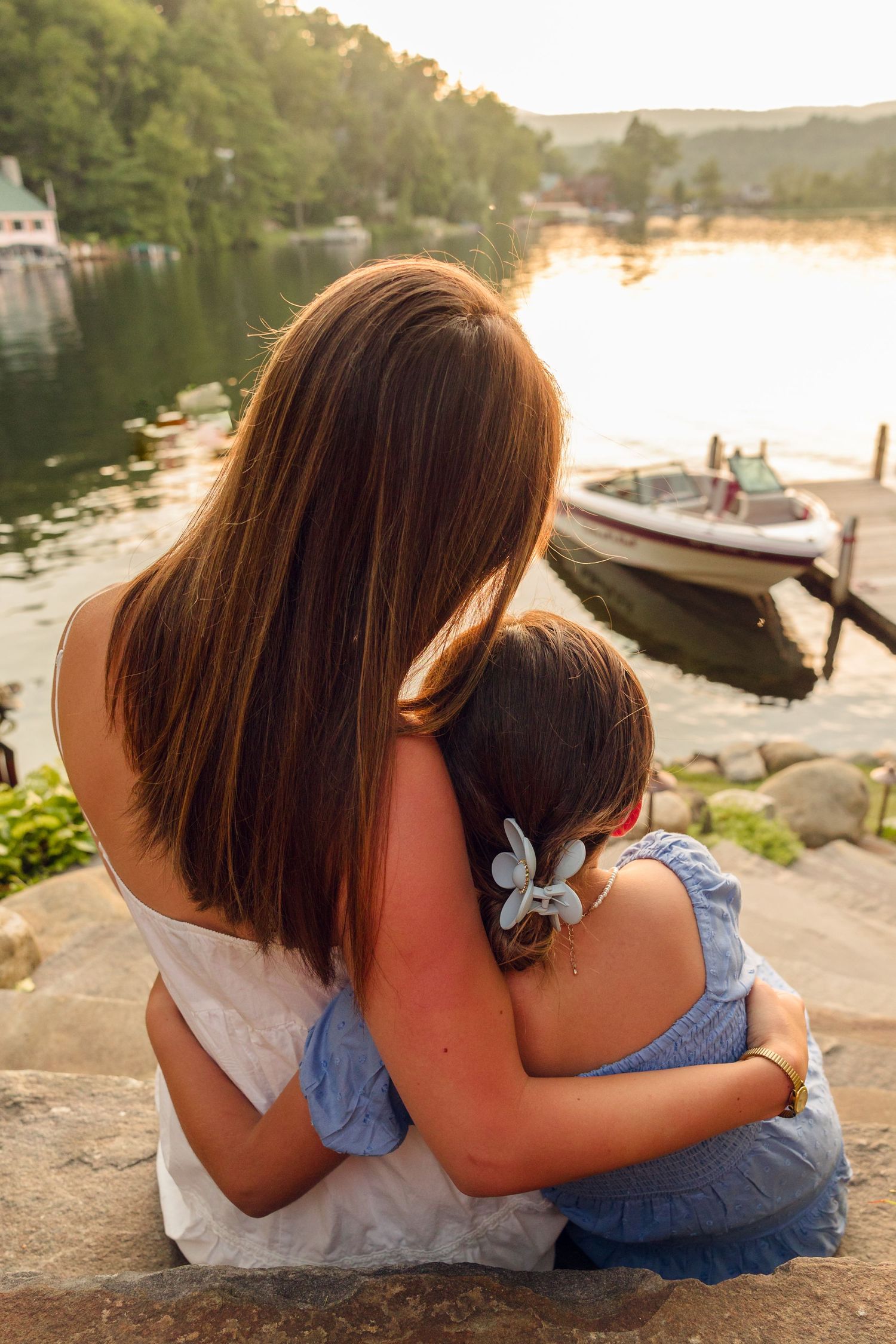 Two people sitting close together on dock with boats and lake in background.