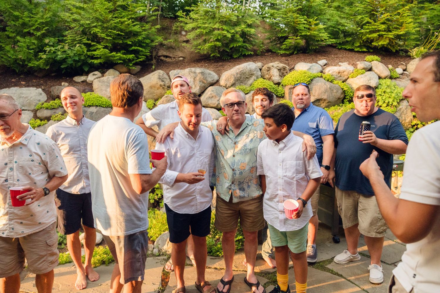 Large group of people gathered outdoors on stone patio holding drinks and socializing.