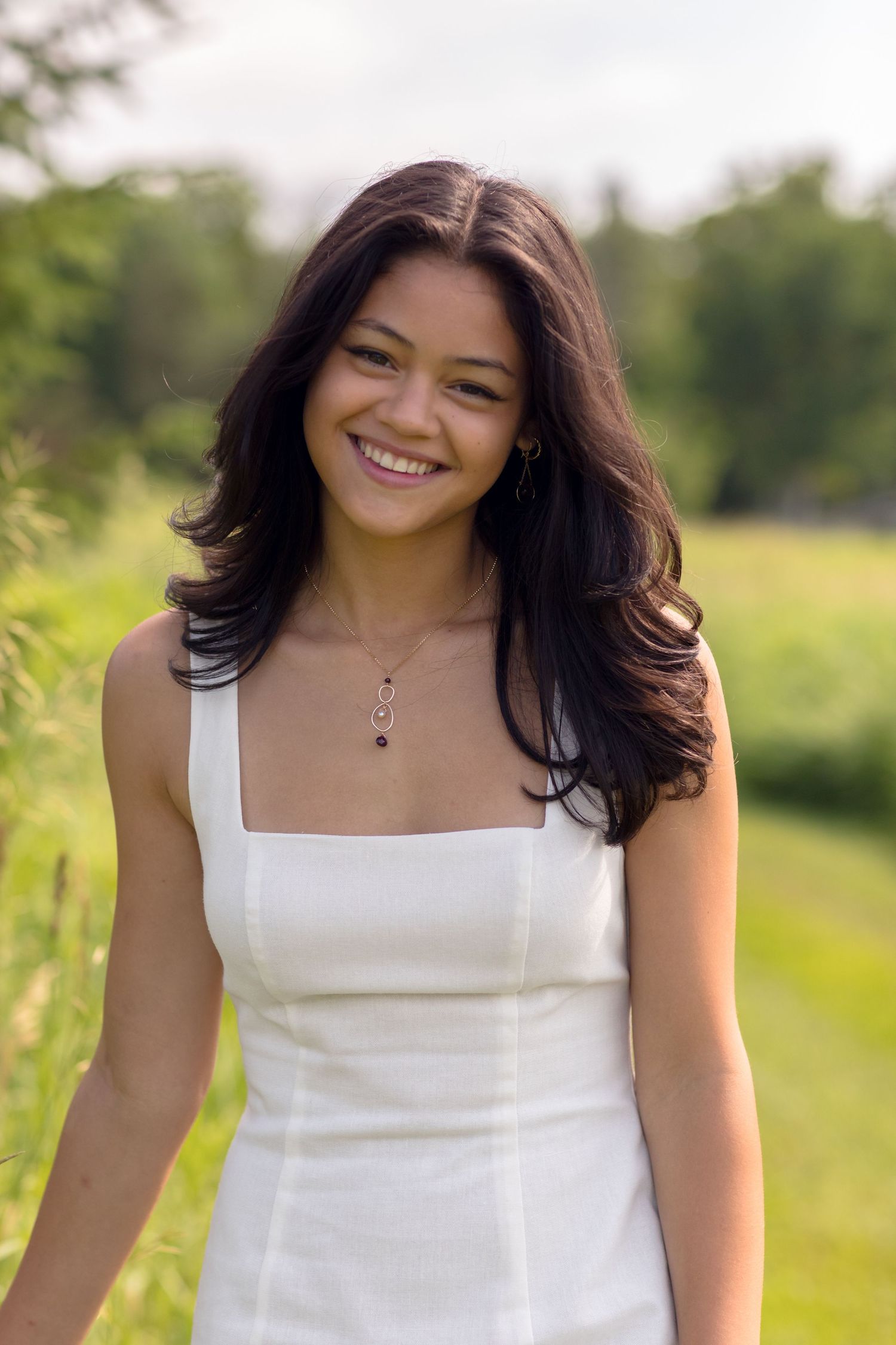 Graduate in white dress smiling while posing in sunny outdoor setting.