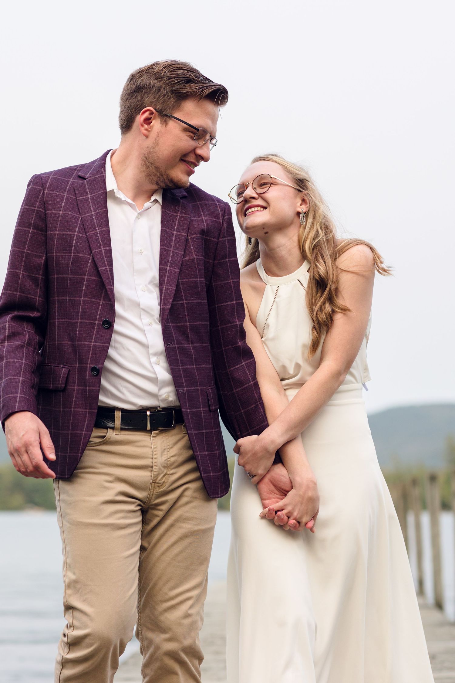 Happy couple holding hands on a dock, man in purple blazer and woman in white dress, smiling at each other.