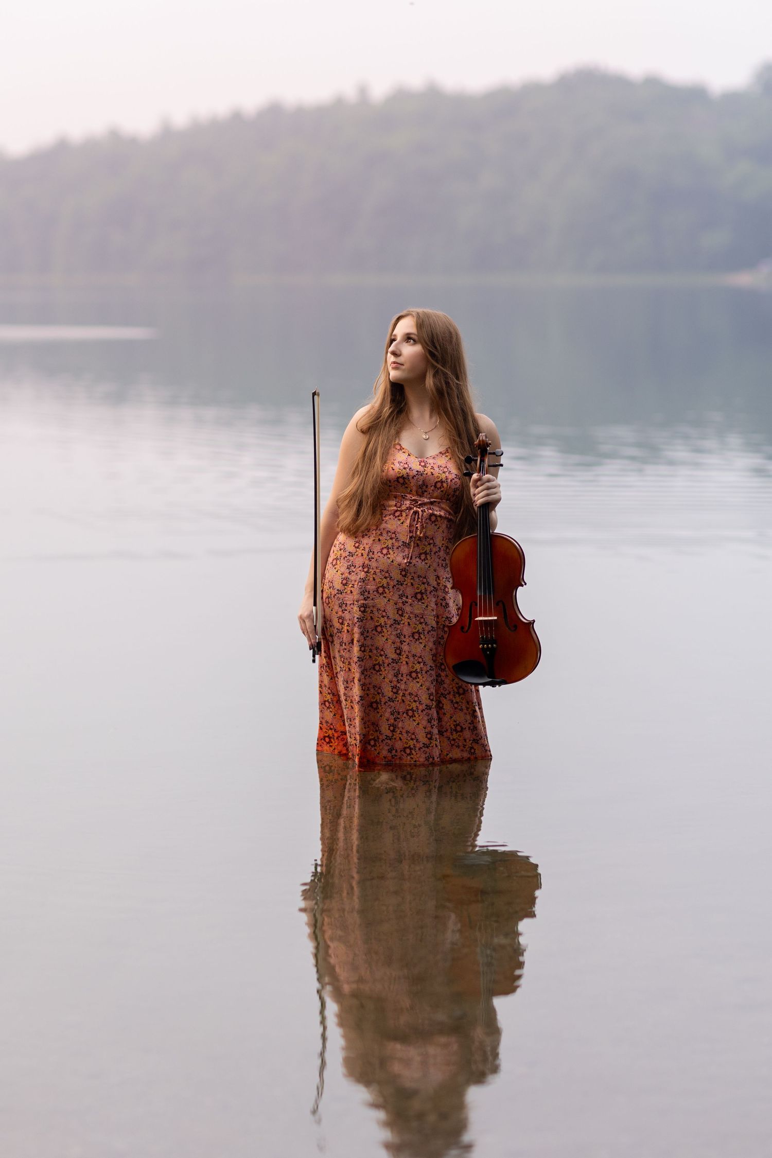 A musician holding a violin stands in shallow lake water during a photoshoot.