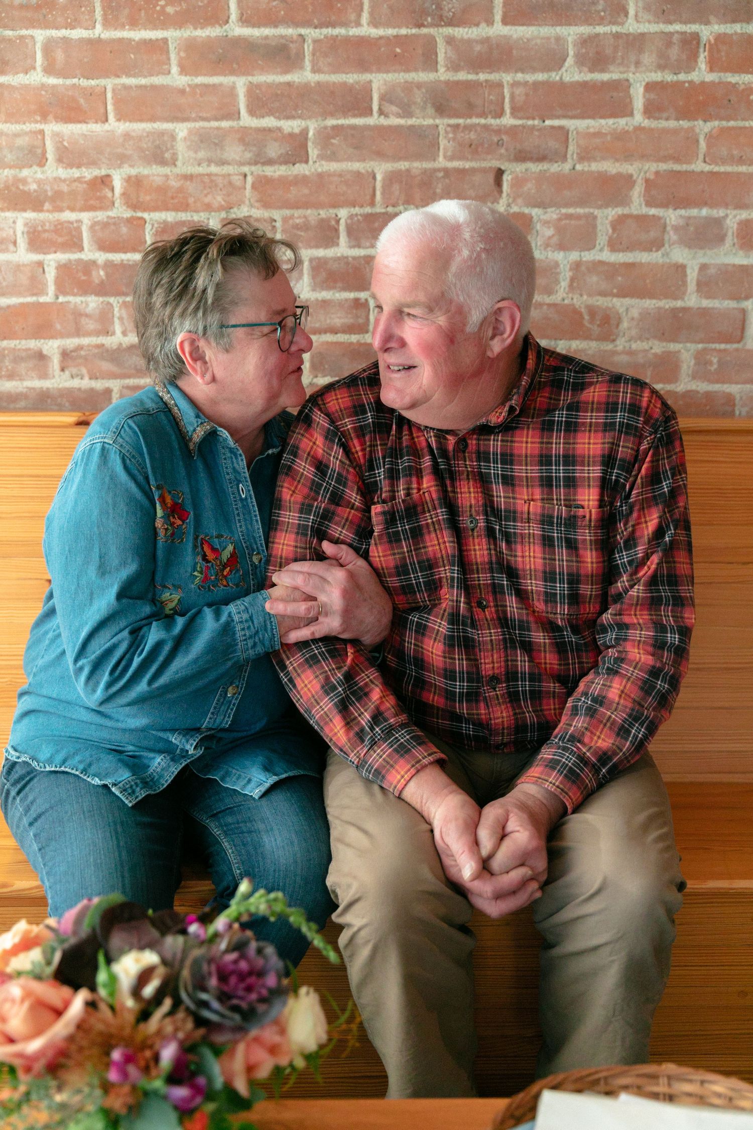 Couple in casual clothing sharing an intimate moment on wooden bench with floral arrangement nearby.