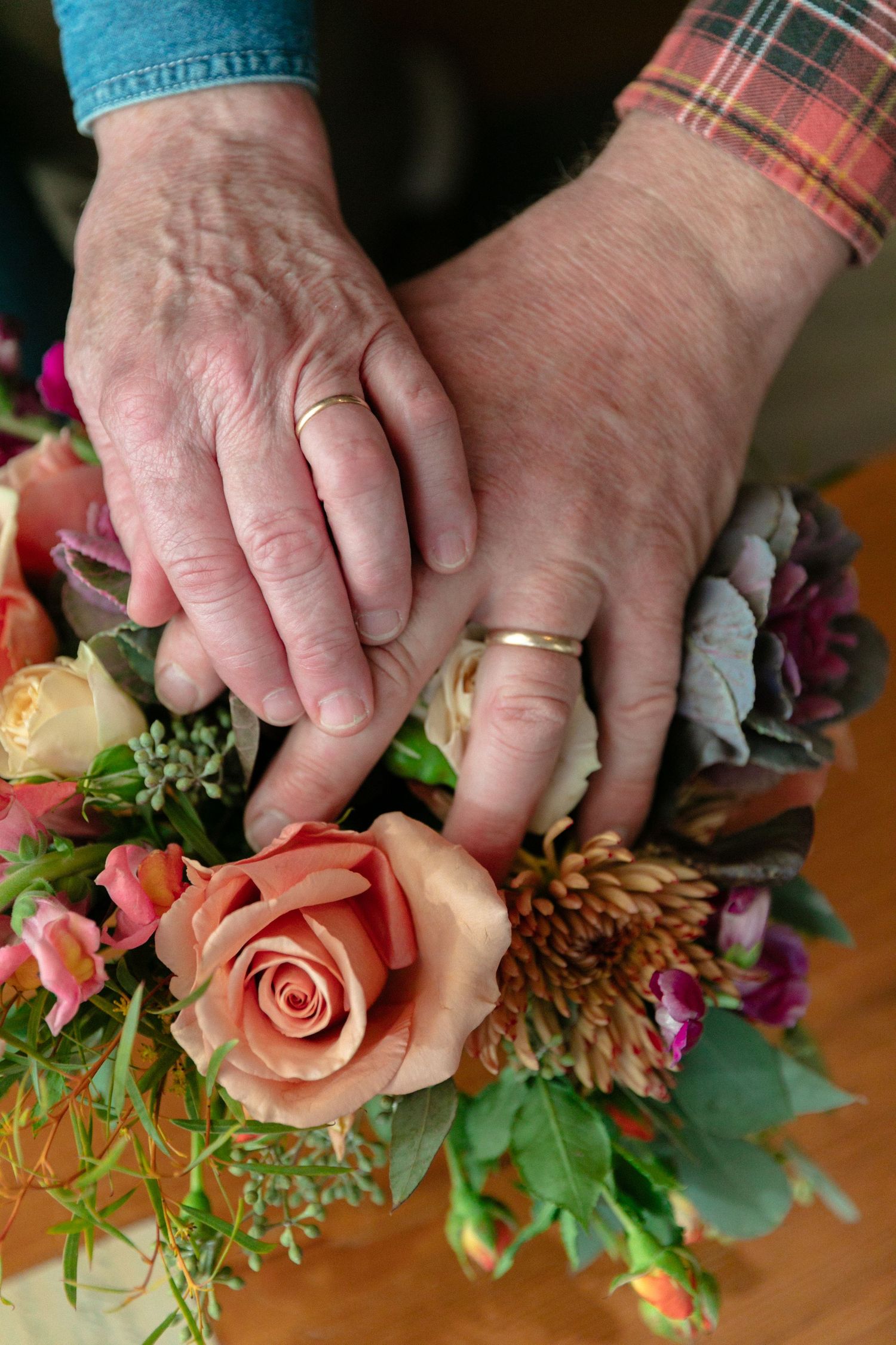 Close up of elderly hands with wedding rings resting on colorful flower arrangement.