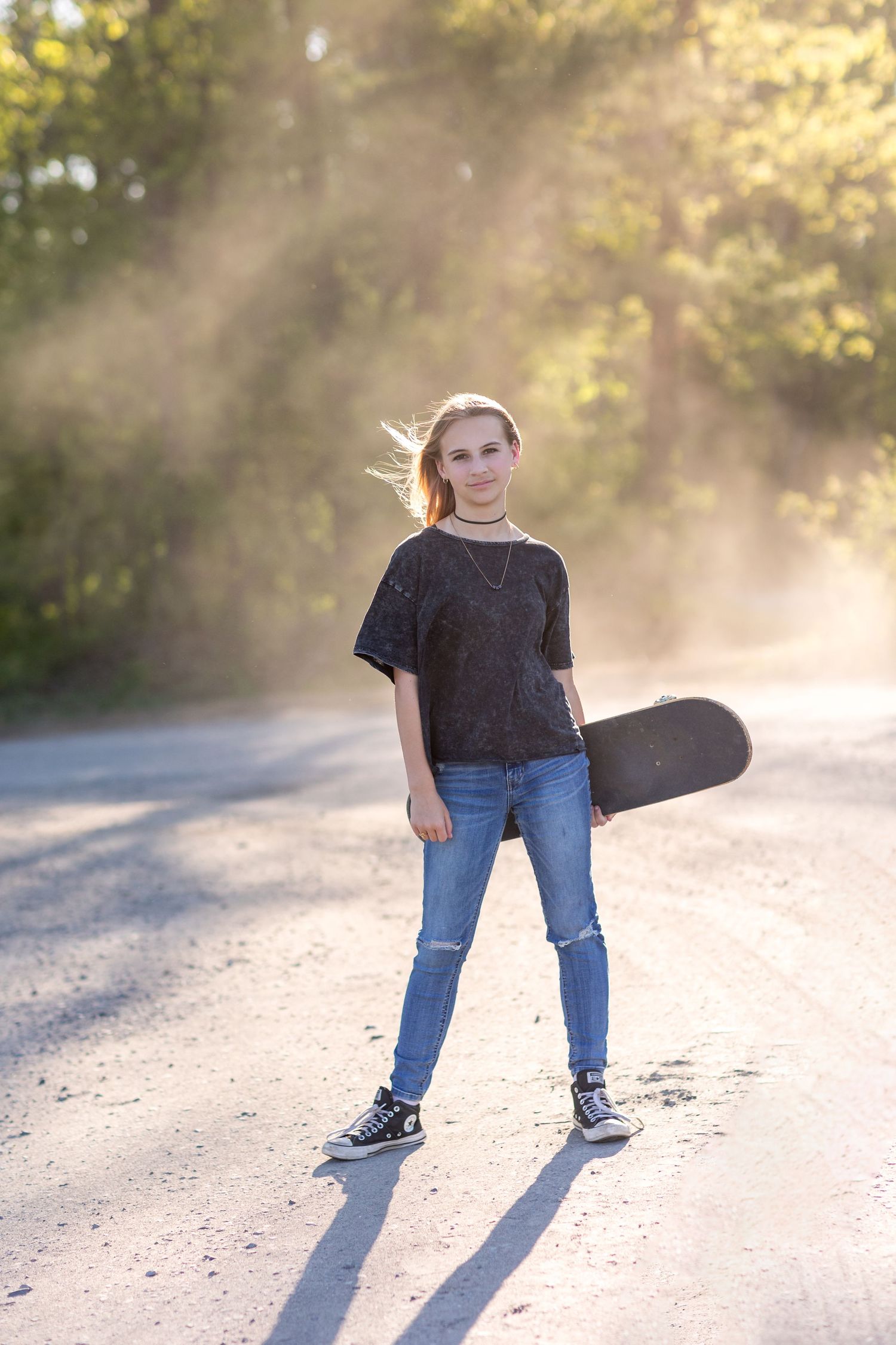 Teen with skateboard stands on misty road in golden afternoon light.