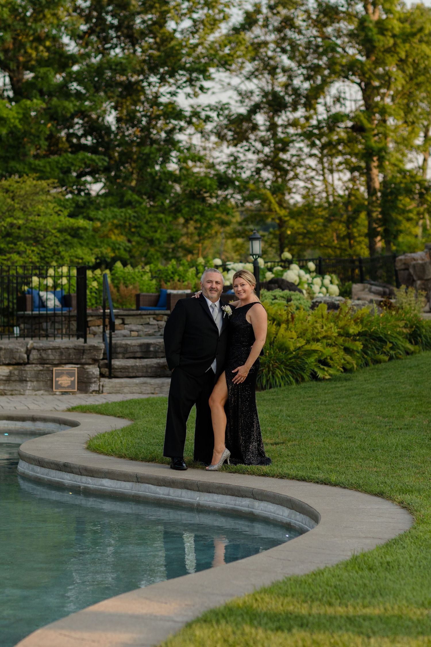 Couple in formal wear poses along a curved pool edge in a landscaped garden setting.