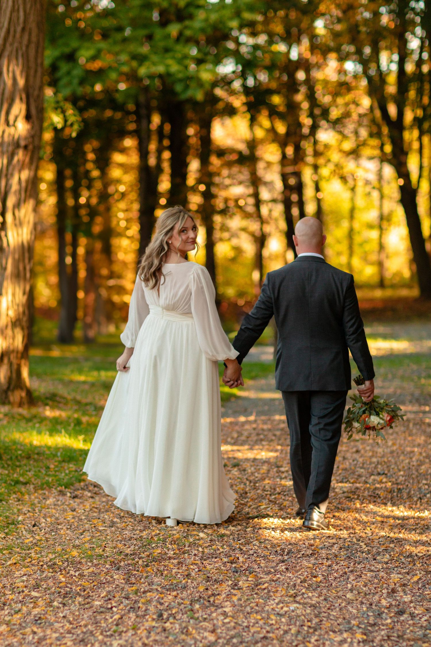 Newlyweds walking hand in hand through a golden autumn forest path.