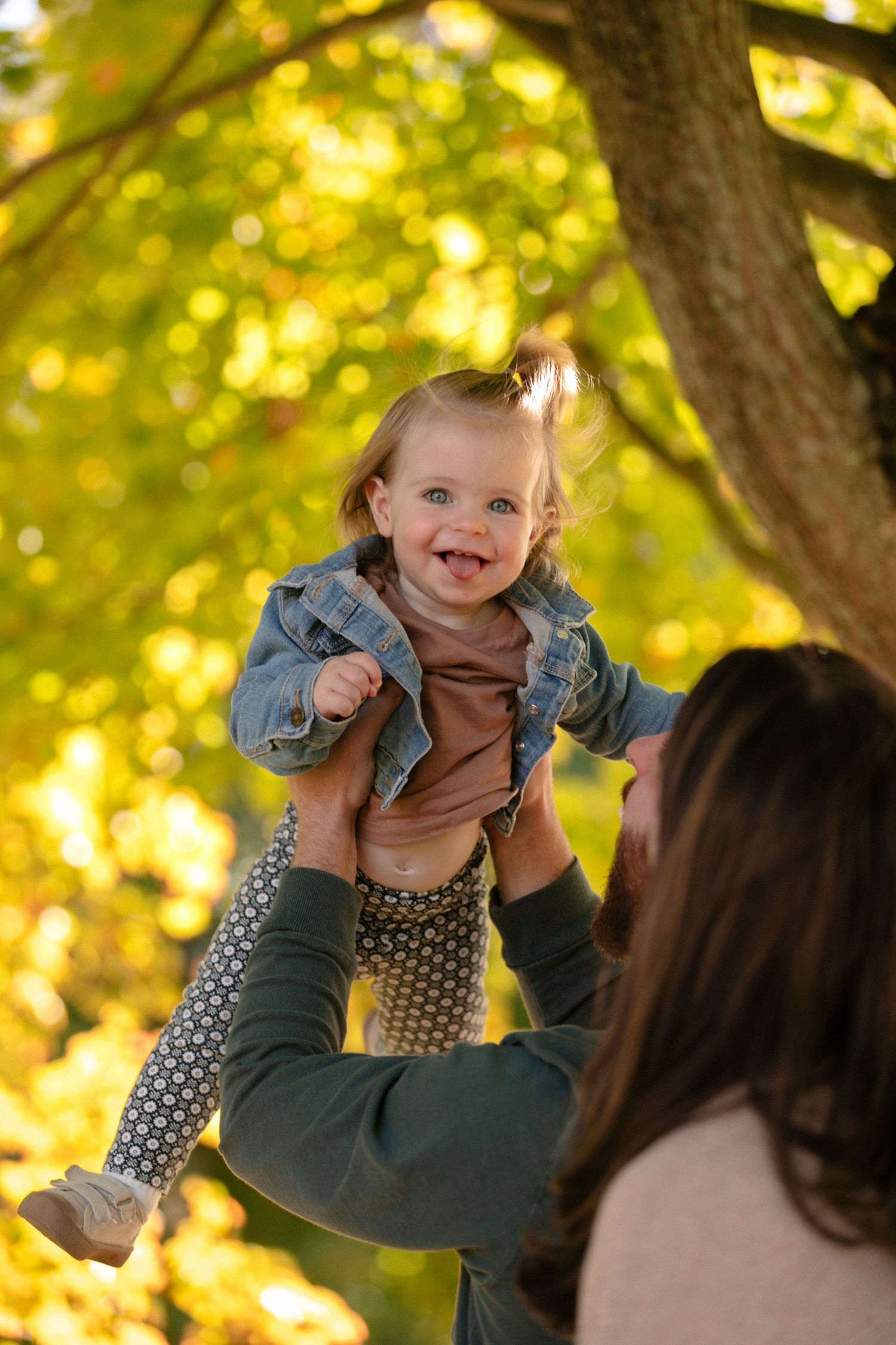 A joyful toddler is lifted high in the air against golden autumn trees.