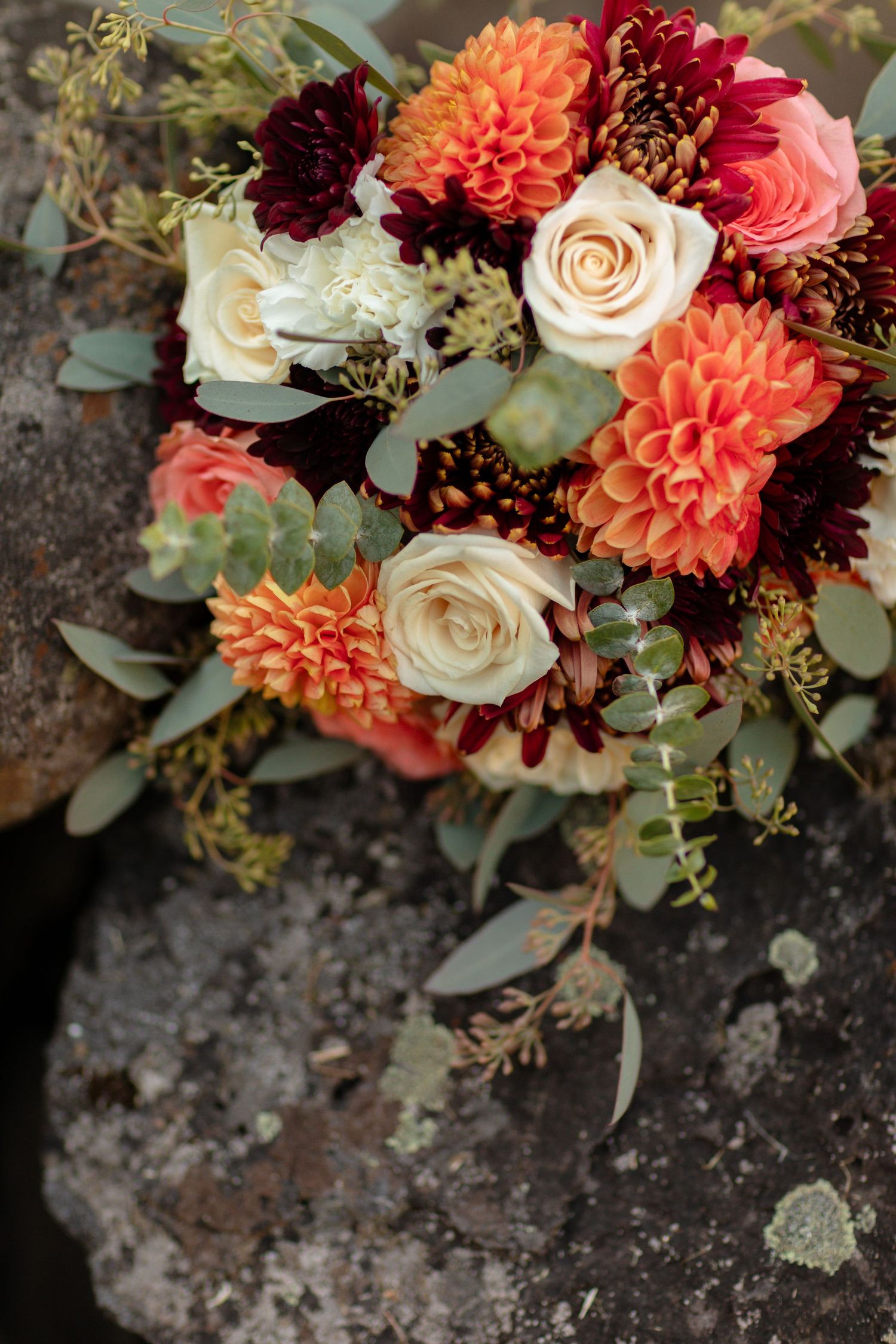 Wedding bouquet with coral dahlias, burgundy flowers, and eucalyptus on stone.
