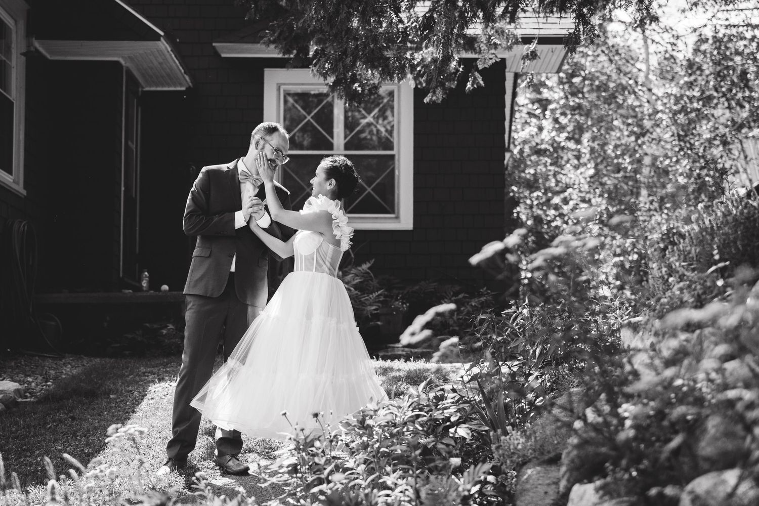 Black and white photo of a bride and groom dancing outdoors near a rustic building surrounded by lush greenery.