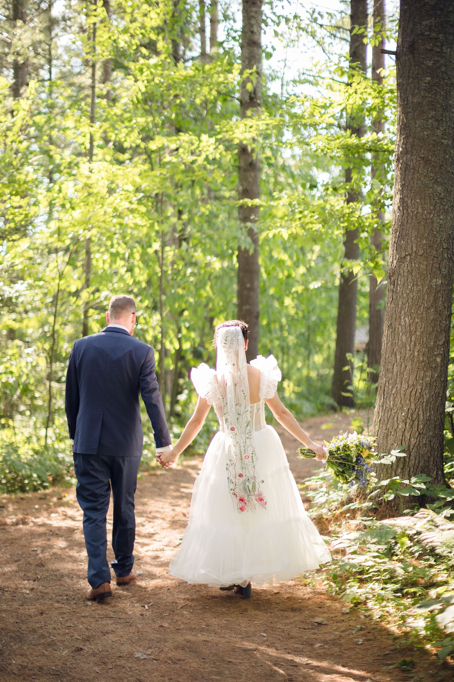 A couple in wedding attire walks through a sunlit forest path in a series of romantic photos.