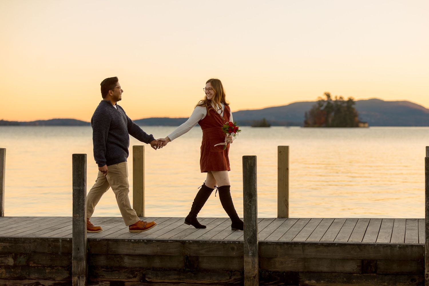 Couple silhouetted against sunset sky on wooden pier by lake.