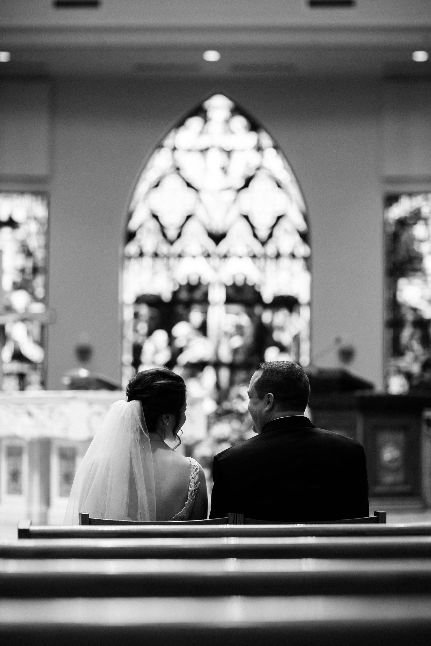A black and white photo of a couple sitting in a church pew beneath stained glass windows.