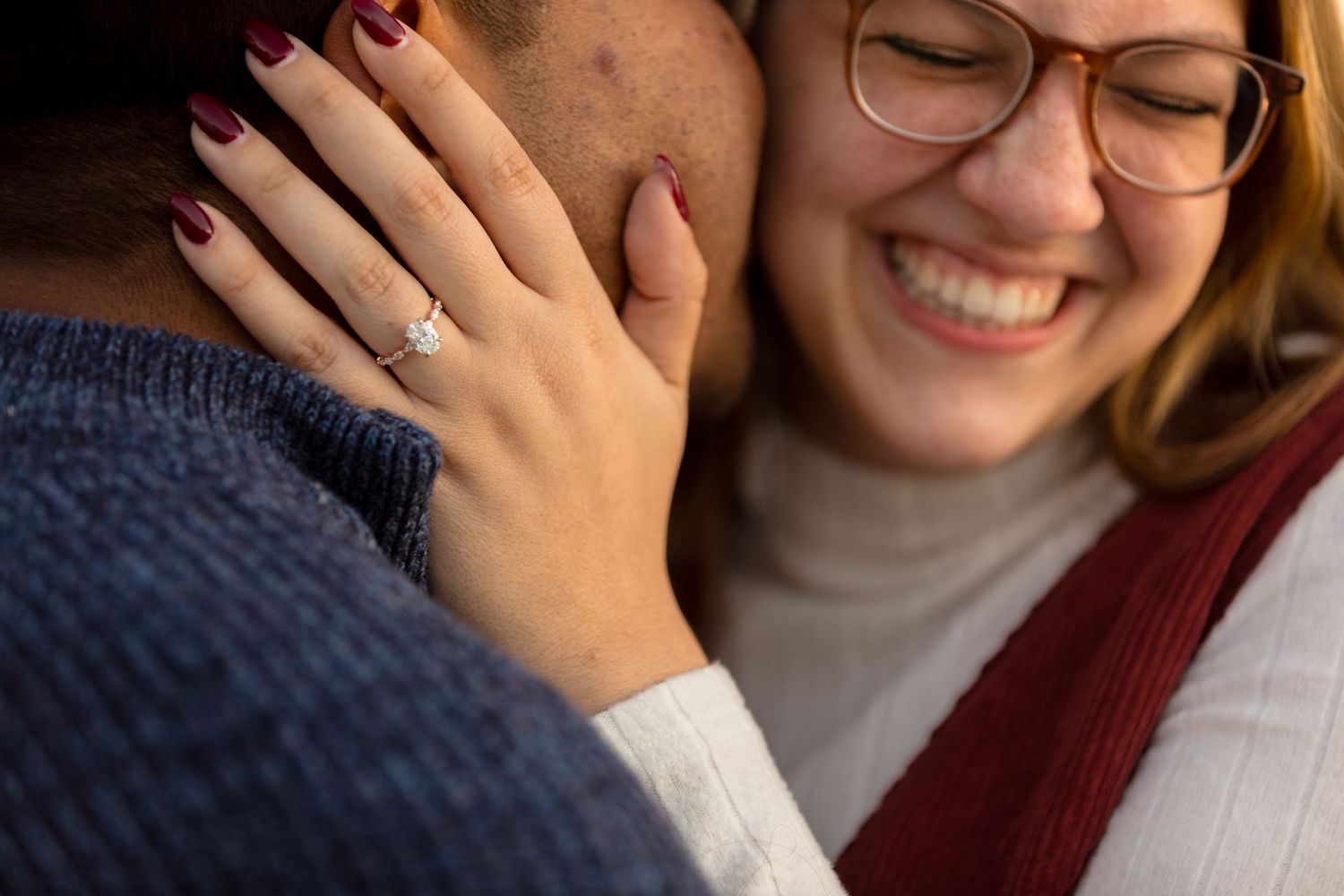 Close up of someone showing off engagement ring while wearing glasses and burgundy sweater.
