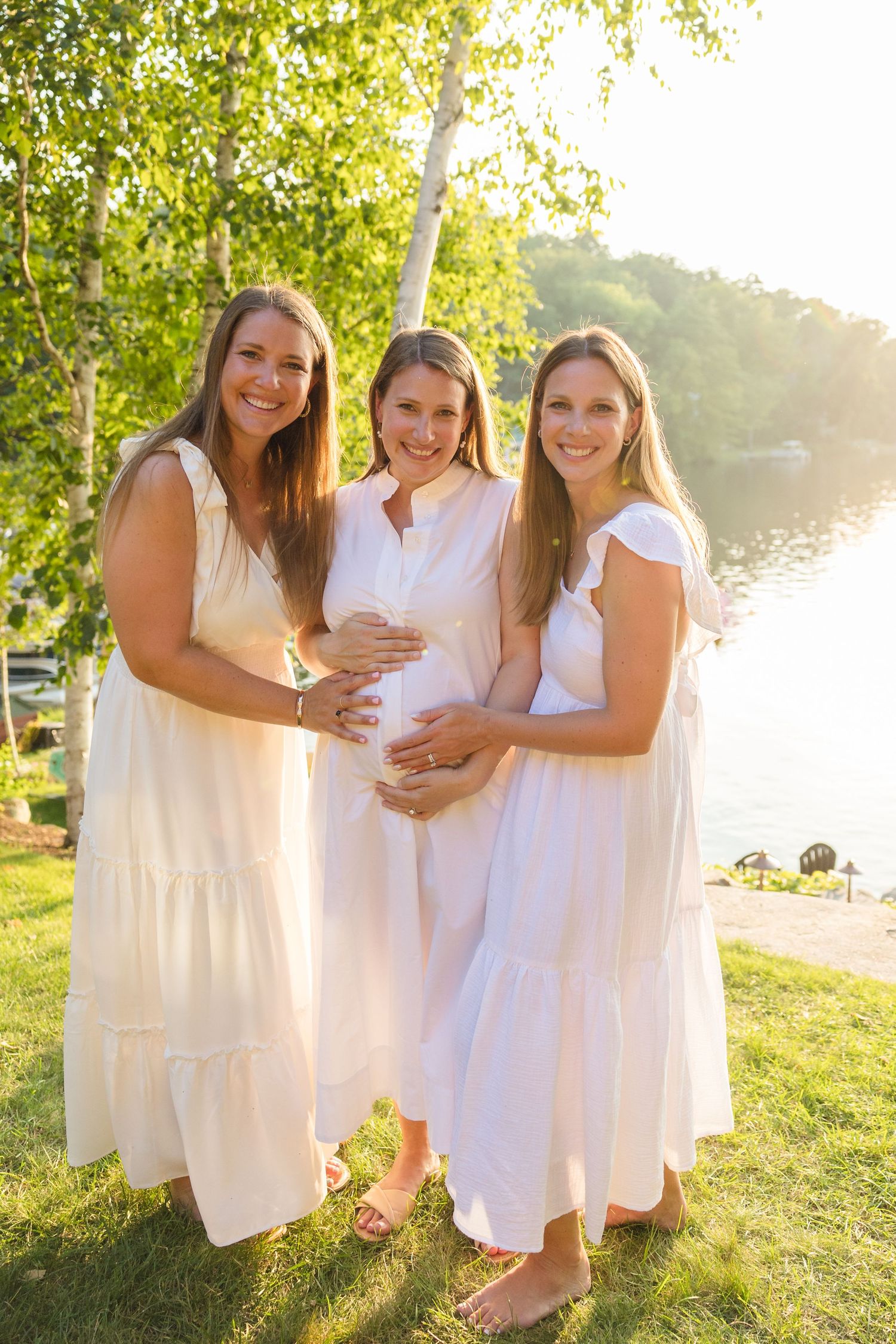 Three women in white dresses pose together by a lake during golden hour.