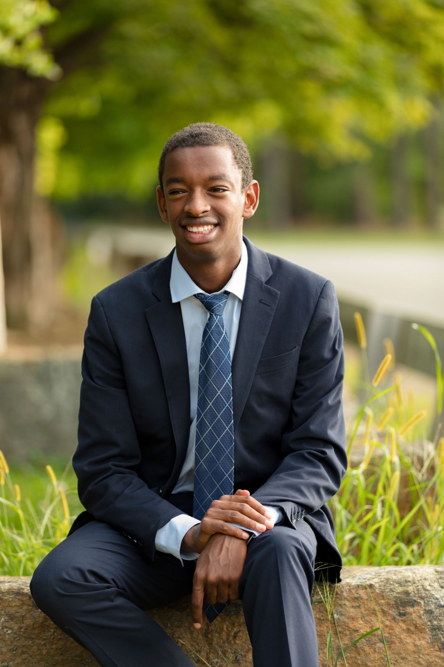 A person in business attire sits casually on a stone wall during an outdoor photo session.