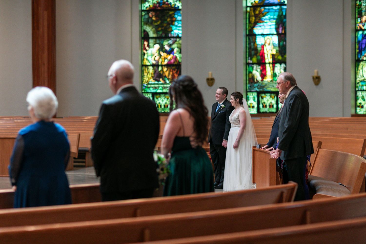 Wedding guests stand in church pews during ceremony with stained glass windows visible.
