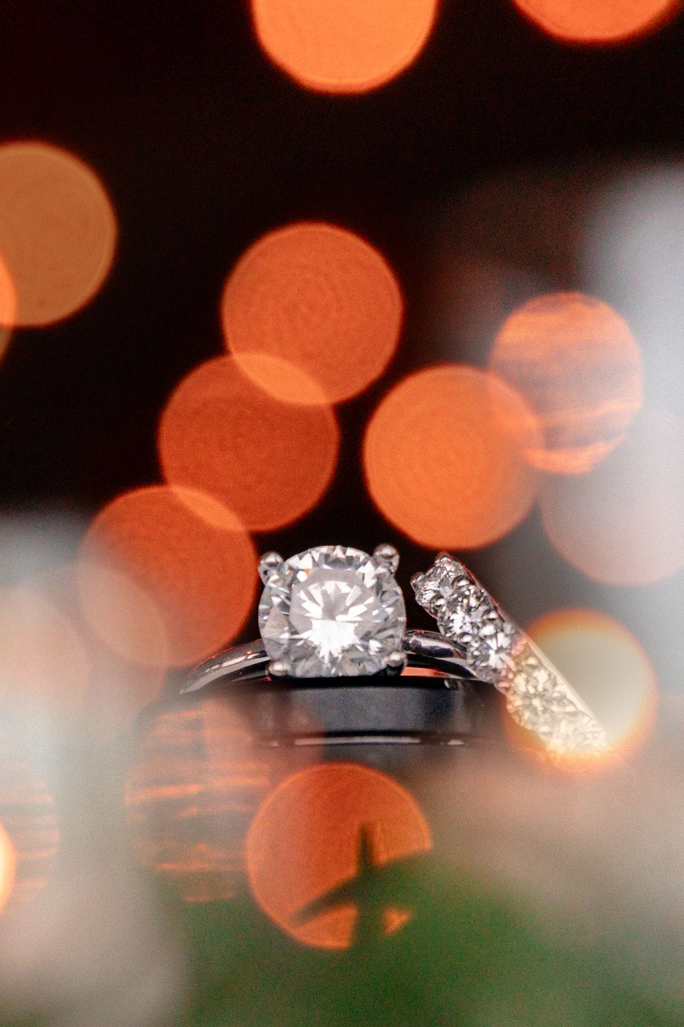 Diamond engagement rings sparkle against a blurred background of orange bokeh lights and green foliage.