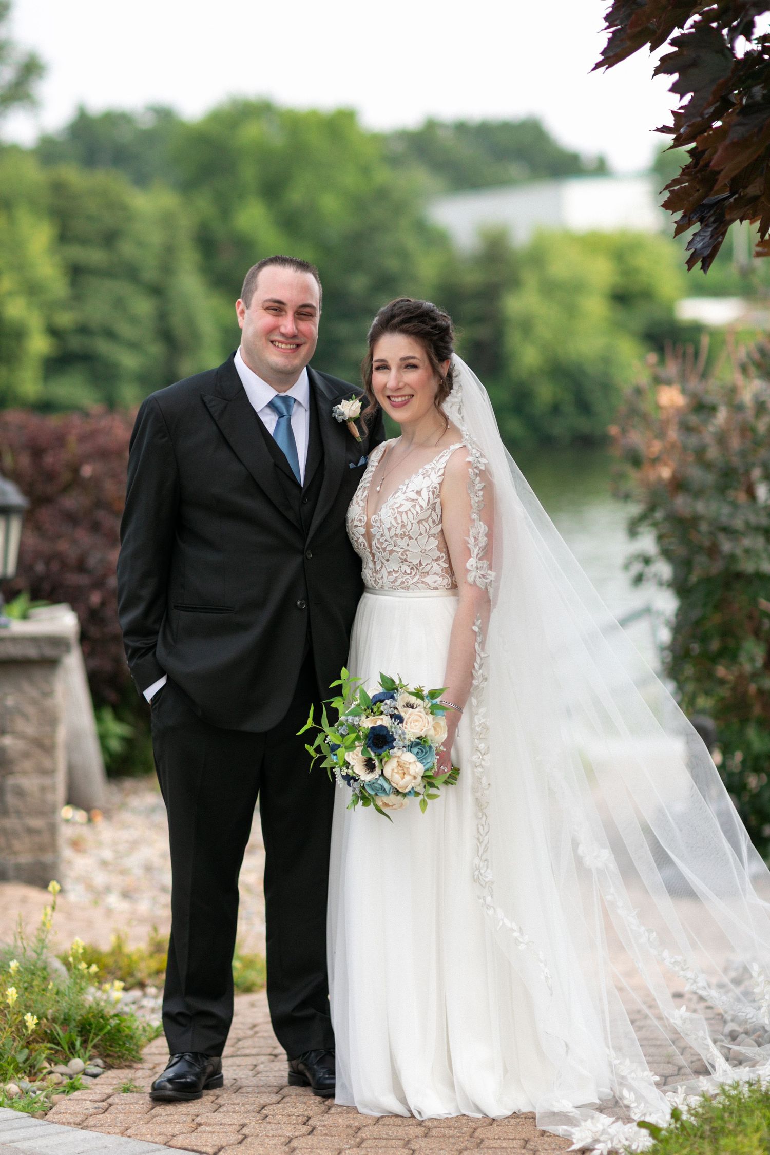 A wedding couple poses outdoors holding a blue and white floral bouquet against a garden backdrop.
