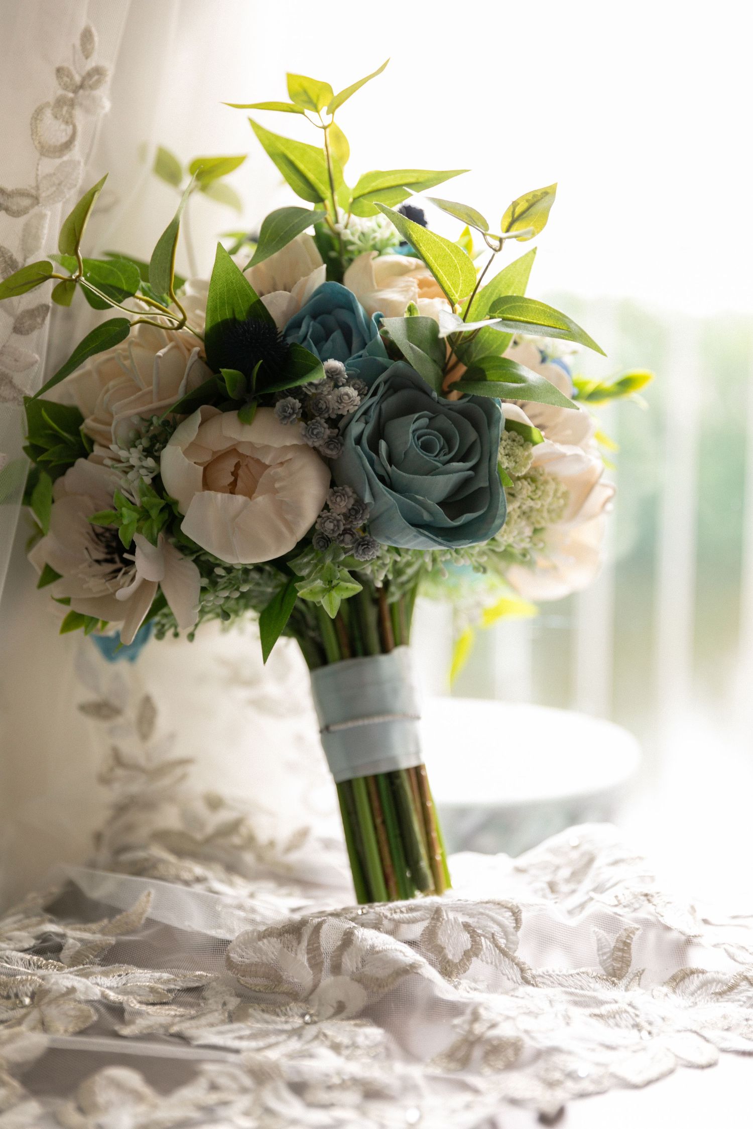 A wedding bouquet with blue and white roses wrapped in white ribbon rests on delicate lace fabric.