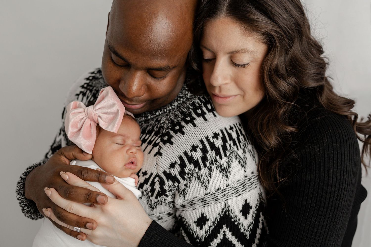Intimate family portrait of parents embracing newborn baby in cozy indoor setting.