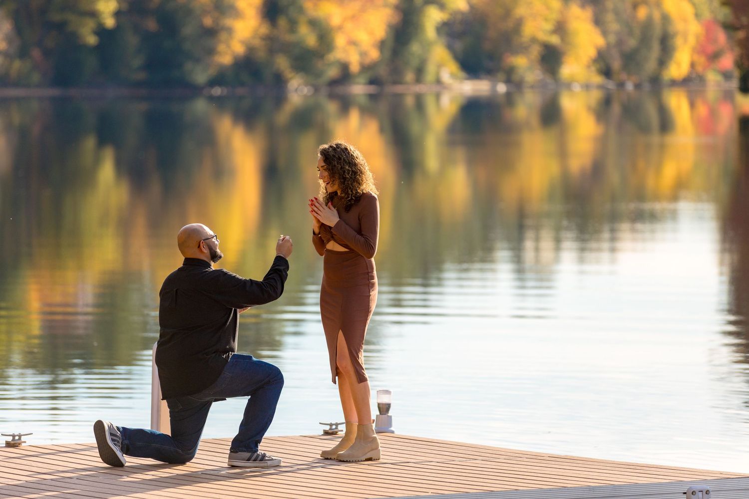 A couple shares a romantic proposal moment on a wooden dock overlooking a serene autumn lake at sunset.