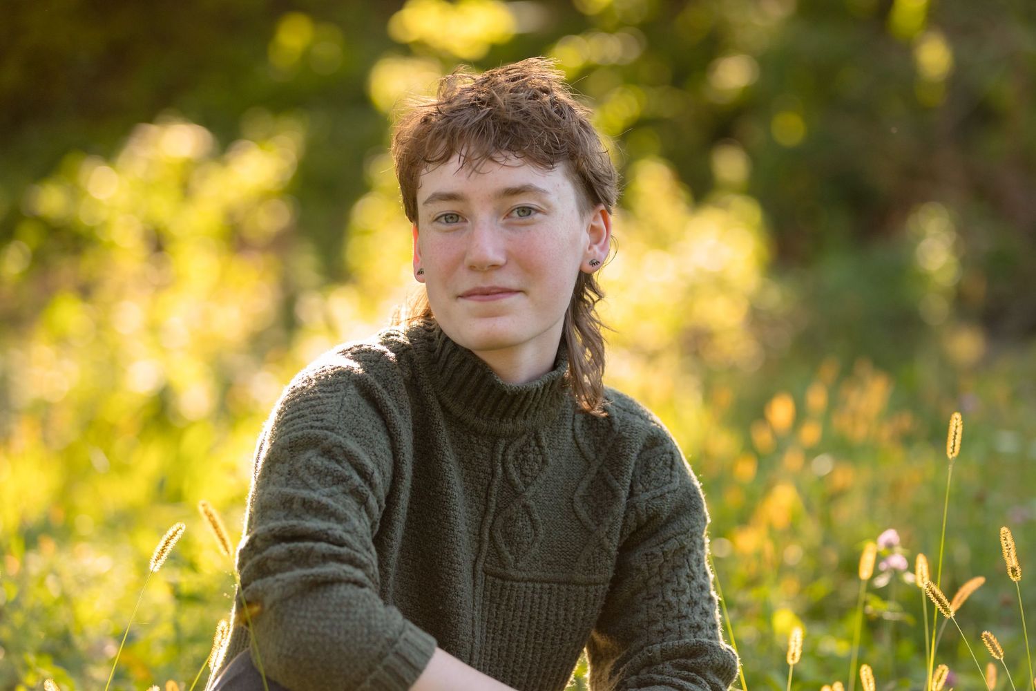 A musician in a green knit sweater sits in golden sunlit meadow.