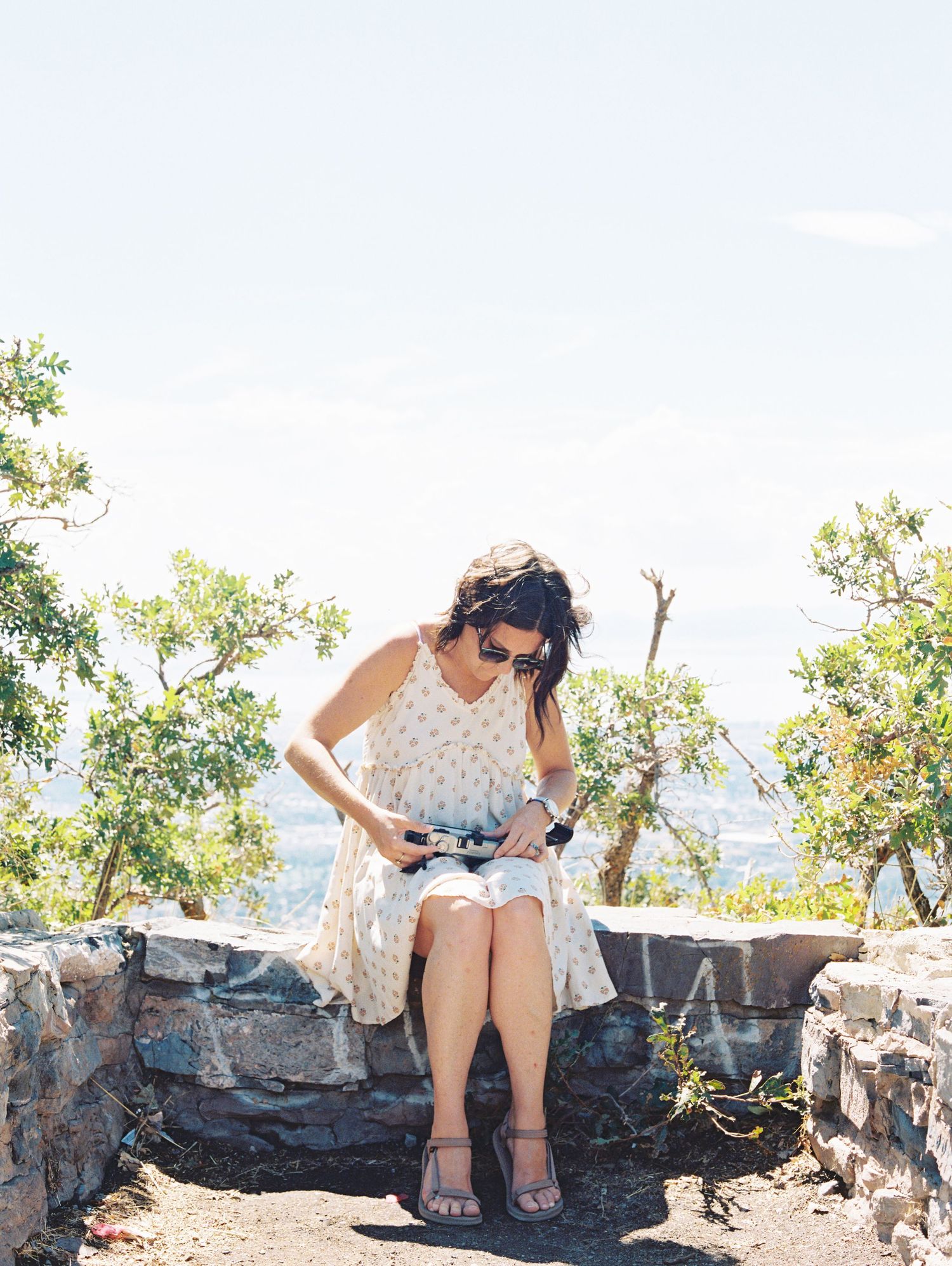 A person sits on a stone wall overlooking a scenic mountain landscape while taking photos.