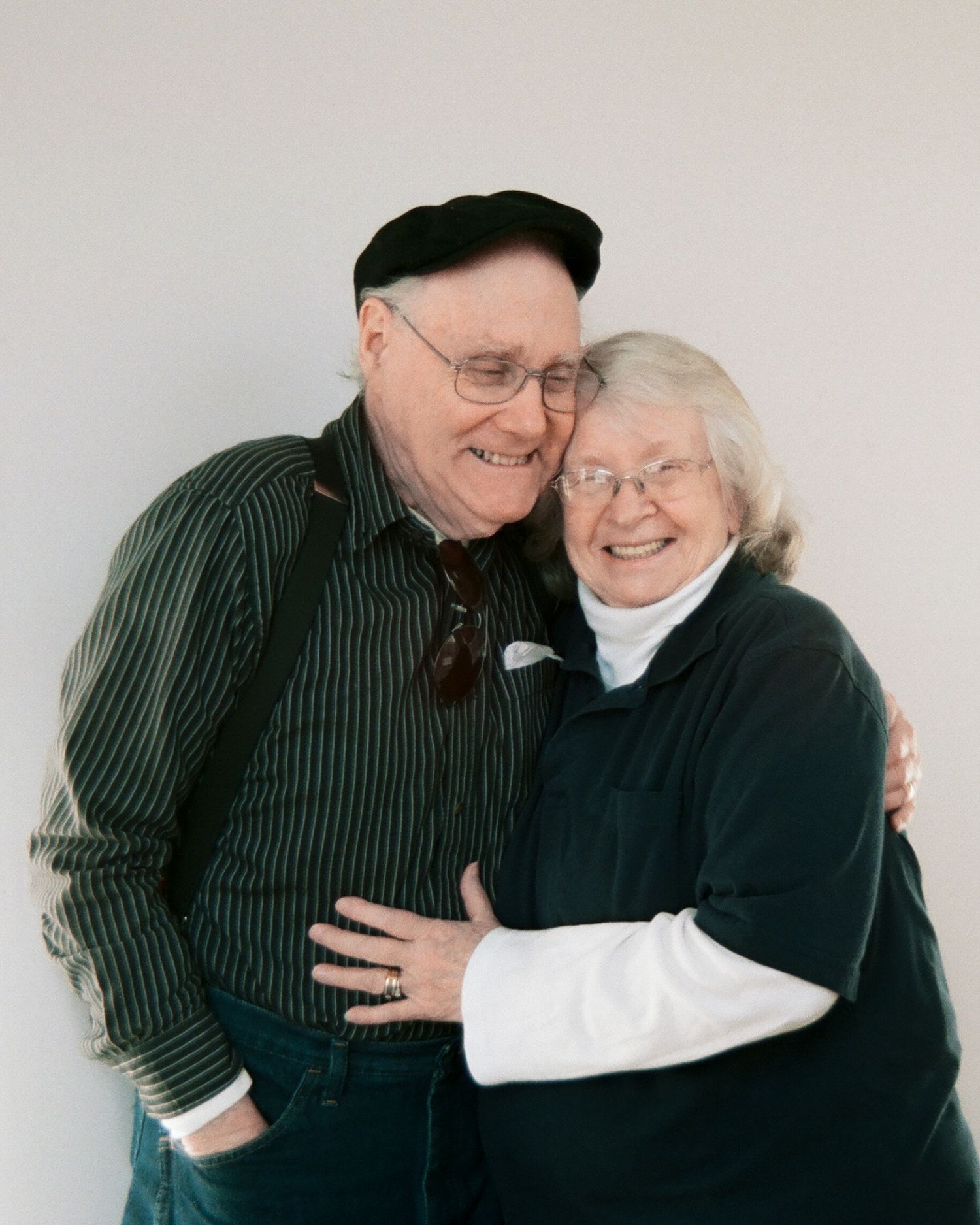 An elderly couple embraces and laughs together, with the man wearing a striped shirt and the woman in a black sweater.