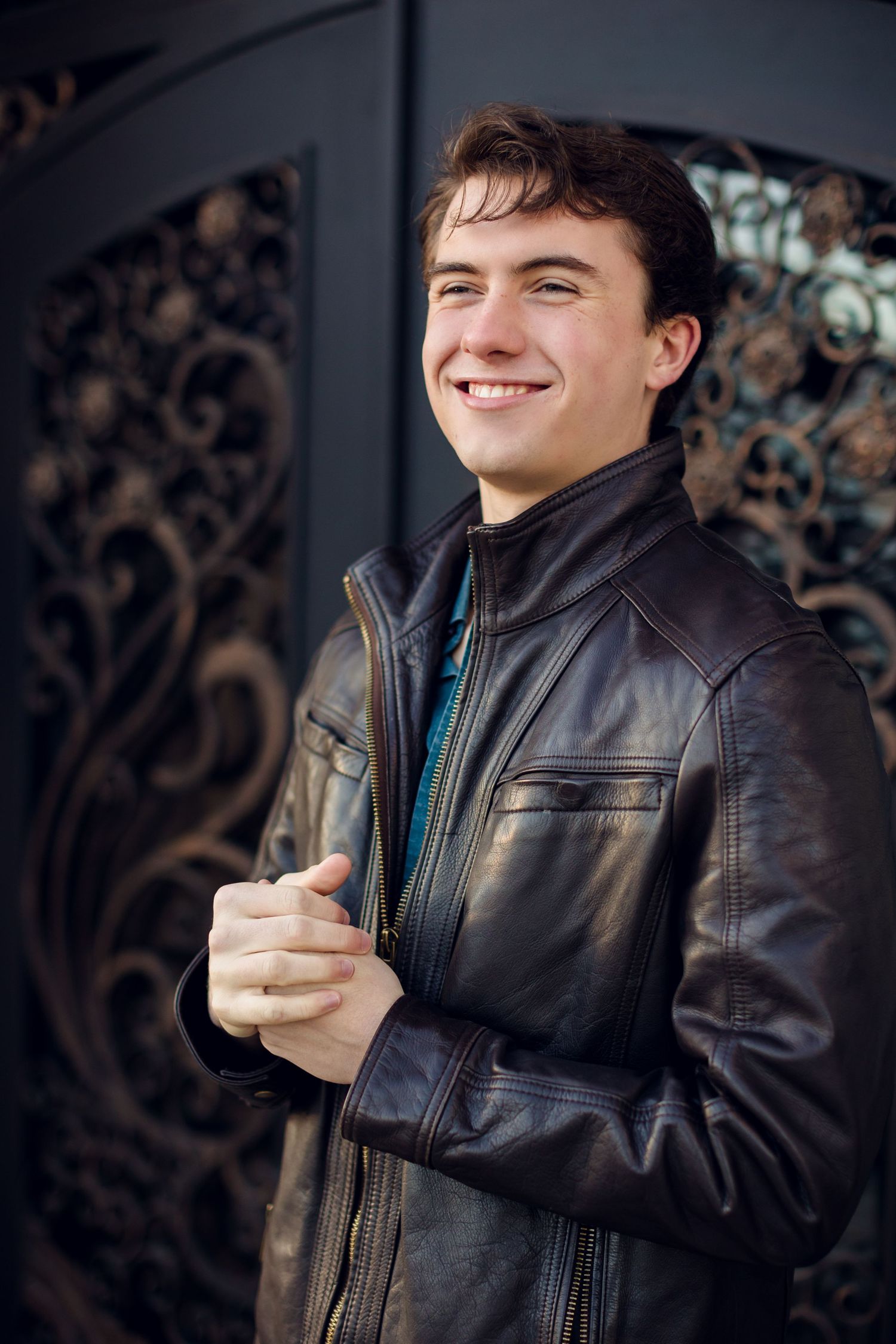 A person in a black leather jacket gestures and smiles while posing against a dark patterned background.