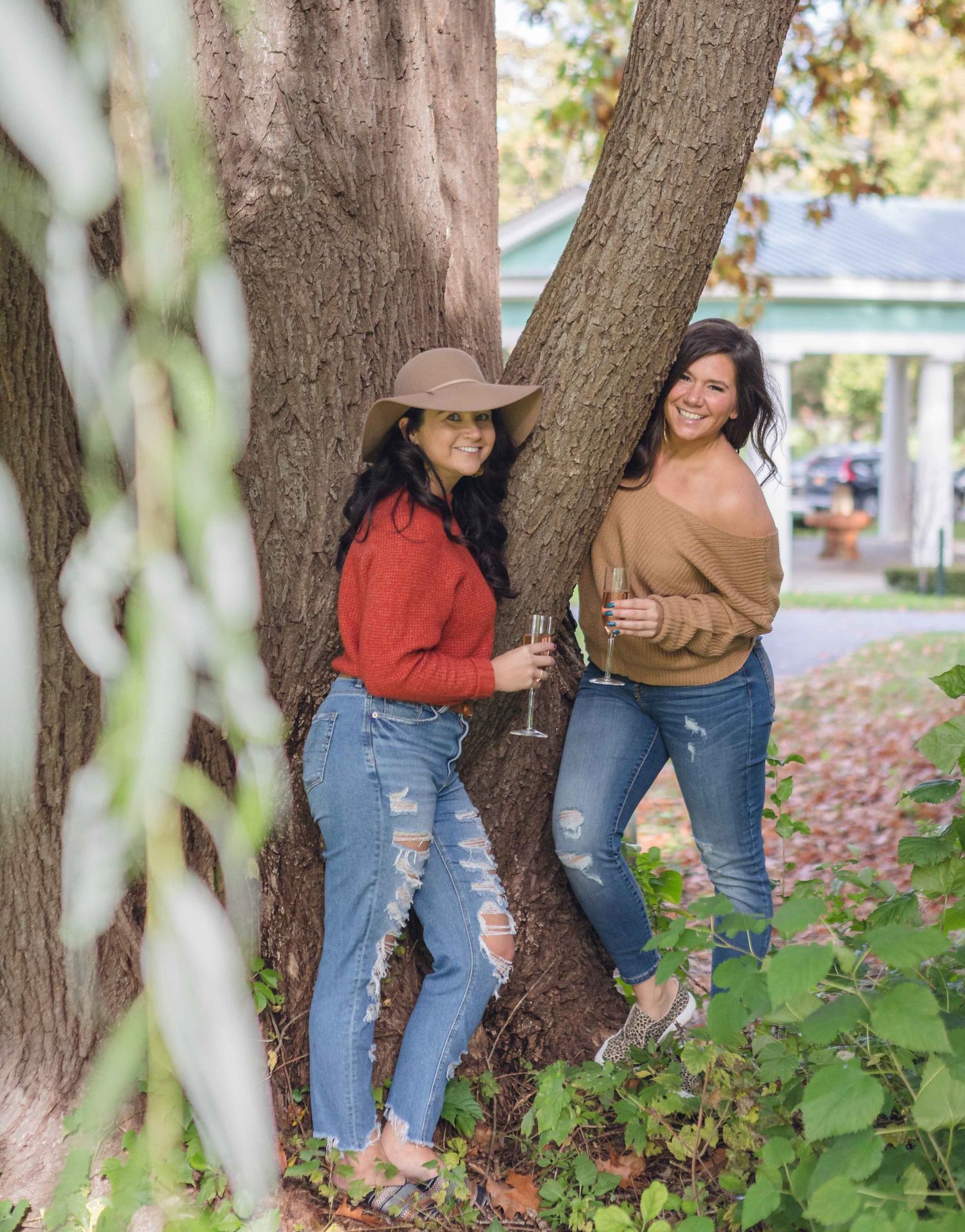 Two women in jeans holding champagne glasses lean against a large tree outdoors in autumn.
