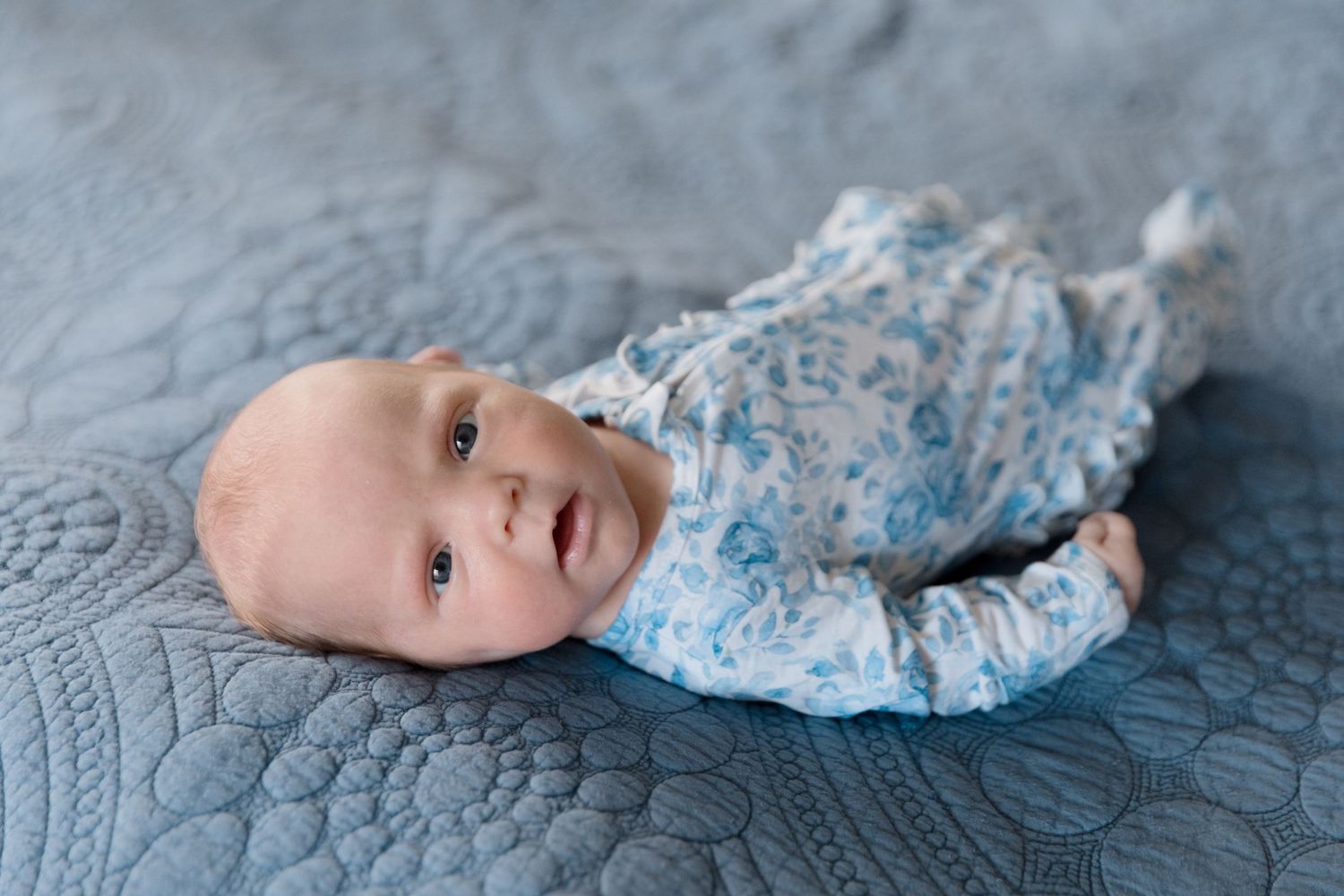 Newborn in blue floral outfit lying on textured grey blanket.