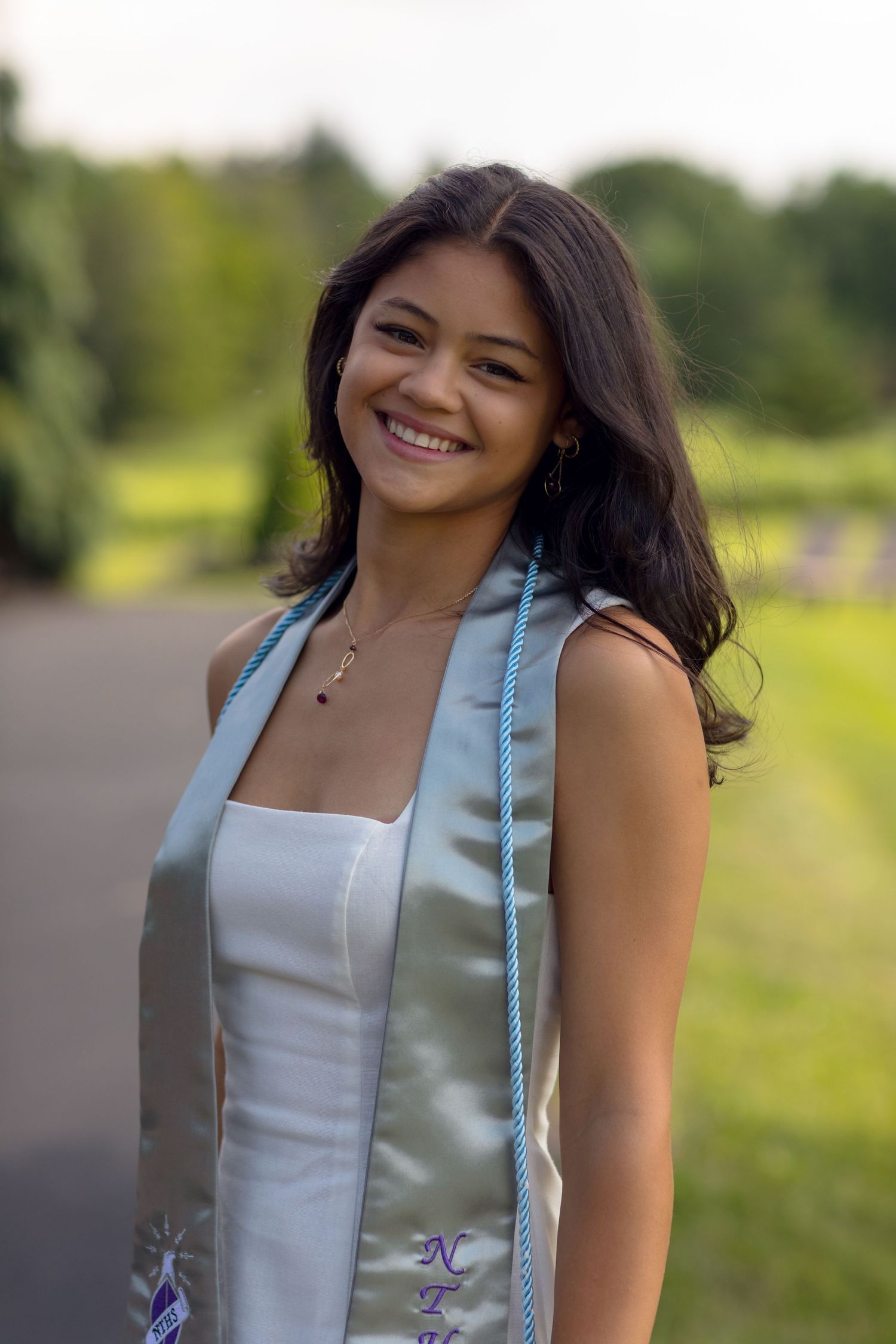Graduate in white dress and light blue stole posing outdoors on a sunny day.