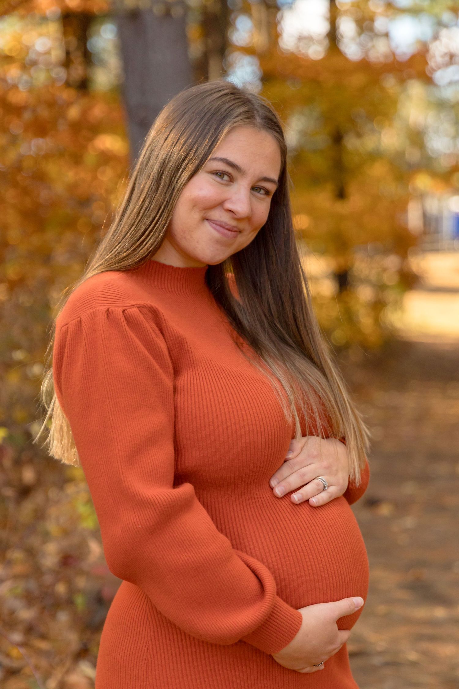 Woman in orange sweater poses with baby bump in autumn forest setting.