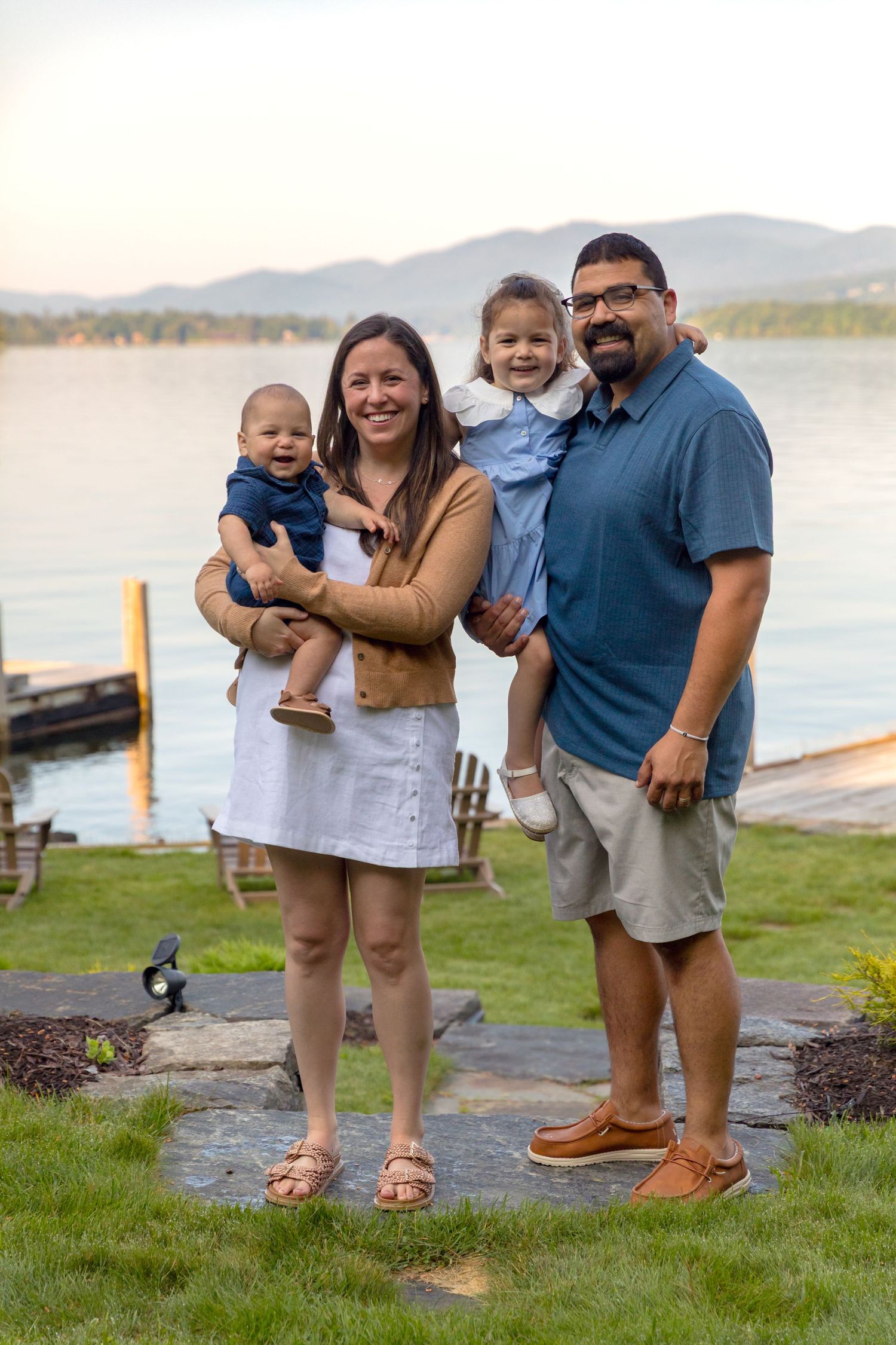 Family poses together on grassy shoreline with mountains and lake in background.
