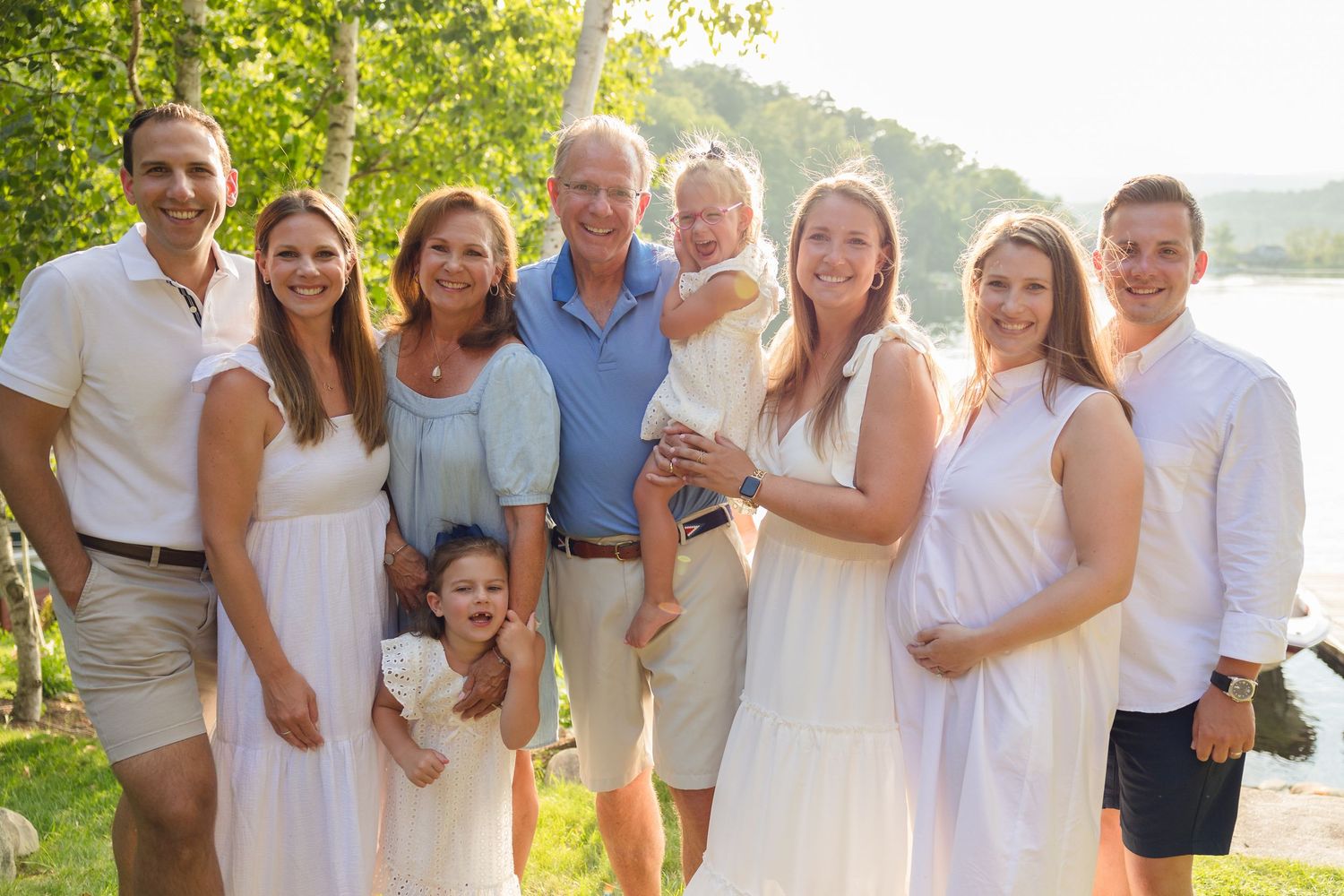 Extended family dressed in white and blue poses together for photos near a lake on a bright summer day.