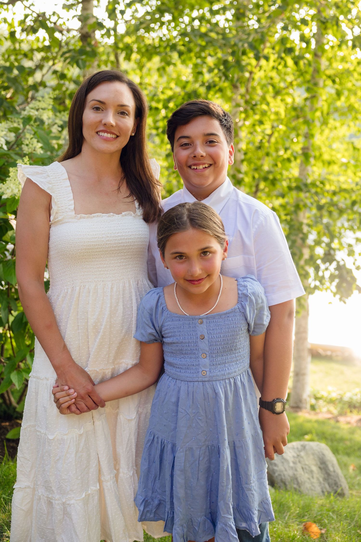 Family of three poses outdoors in summer attire against a natural green backdrop.