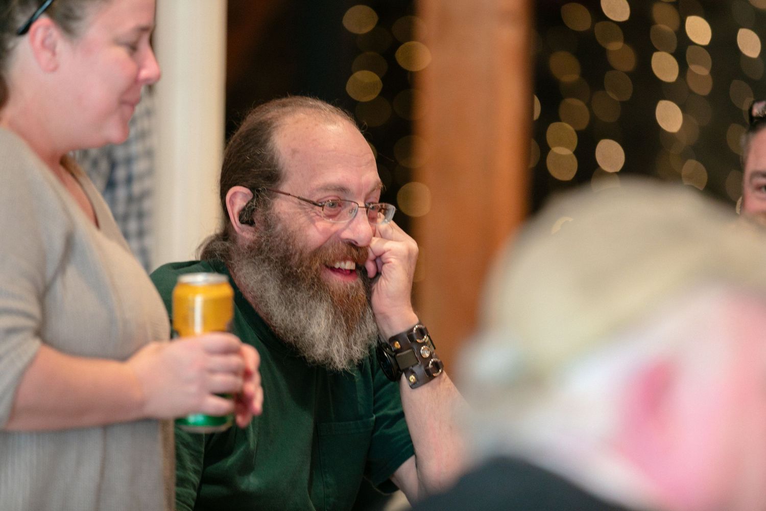 A bearded person in glasses and green shirt laughs during a social gathering.