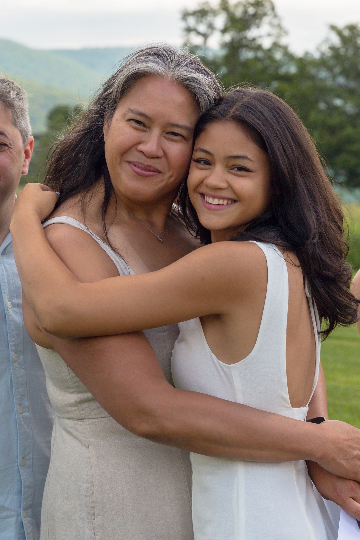 Two people in white tops share a joyful embrace while posing outdoors in a grassy field with mountains in the background.