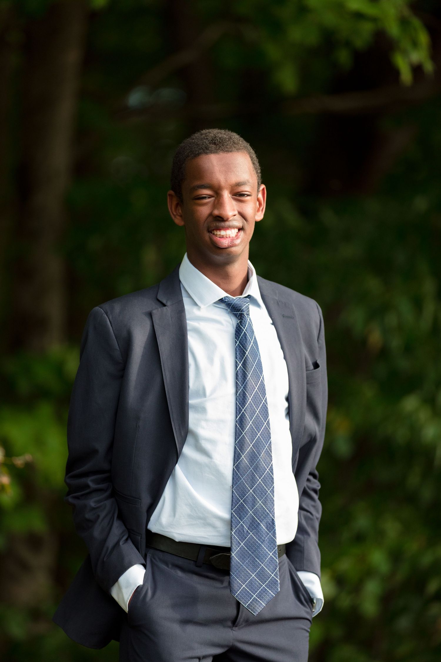 Professional portrait of someone in a dark suit and blue tie smiling against a natural green background.
