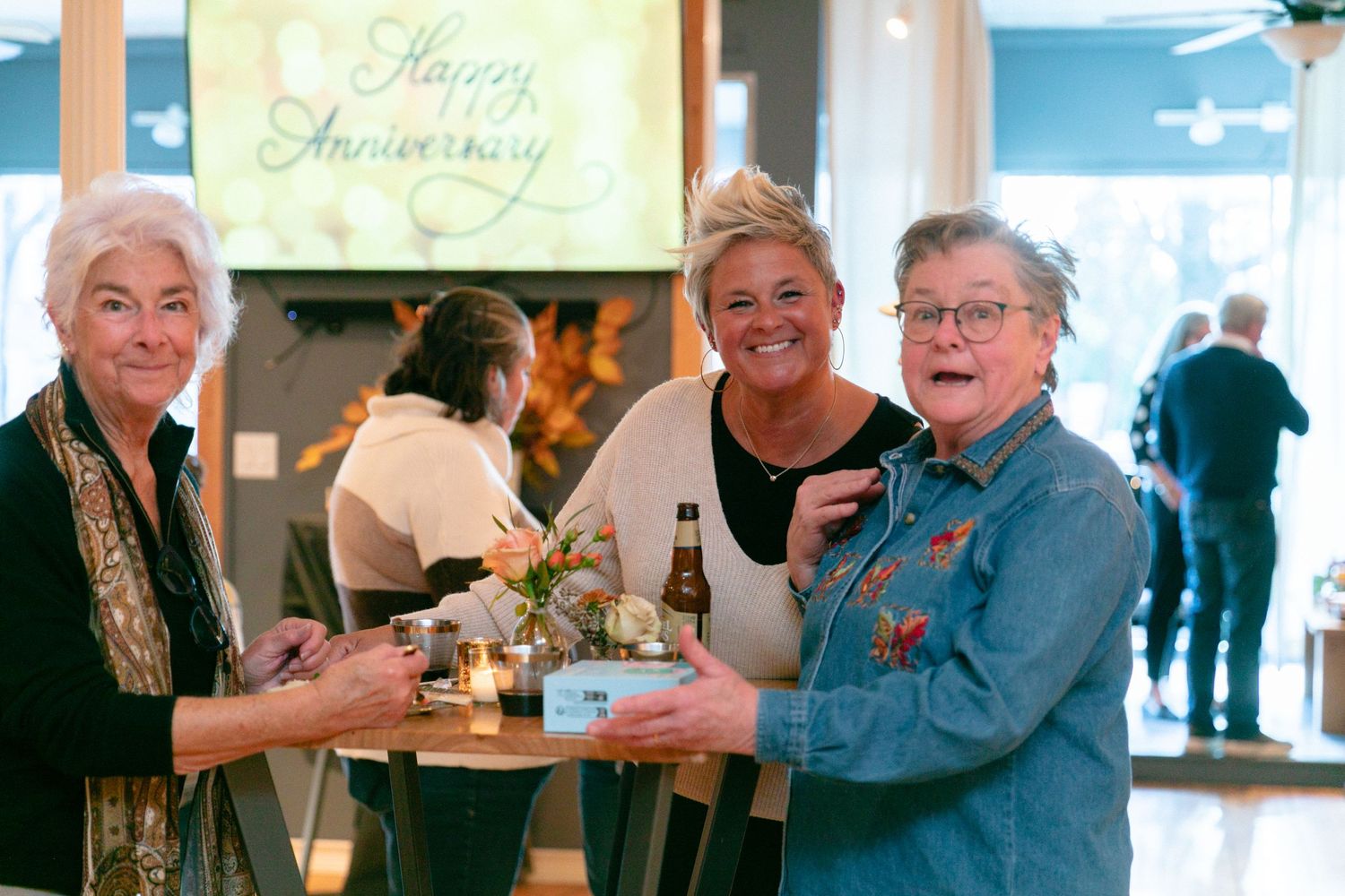 Three seniors share drinks and conversation at a celebration with a Happy Anniversary banner in the background.