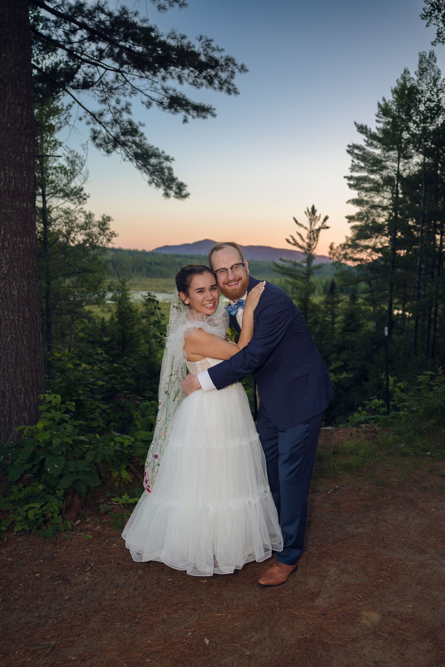 A couple in wedding attire shares a romantic moment at sunset with pine trees and mountains in the background.