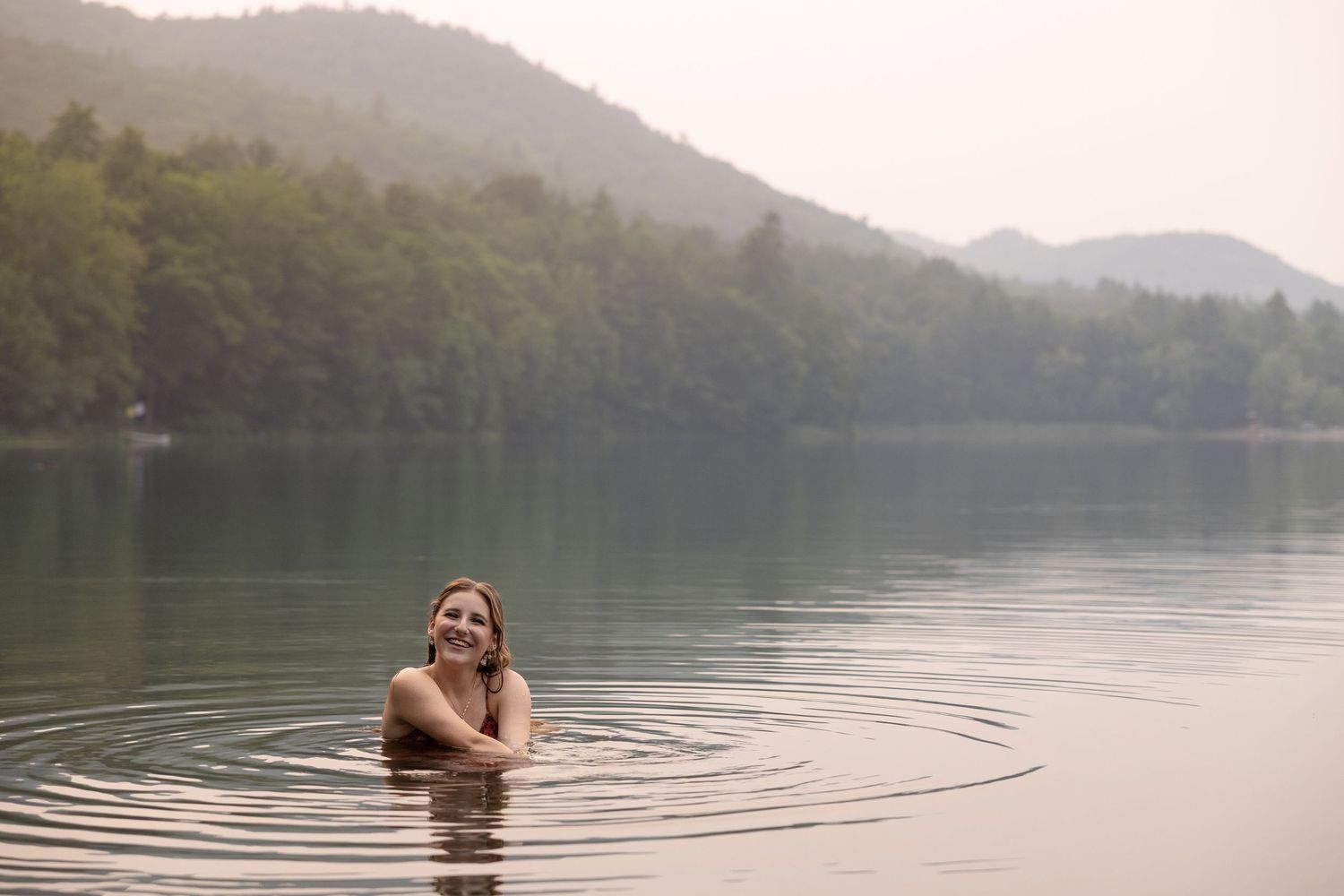 A serene lake scene with ripples in the water surrounded by forested mountains in soft morning light.