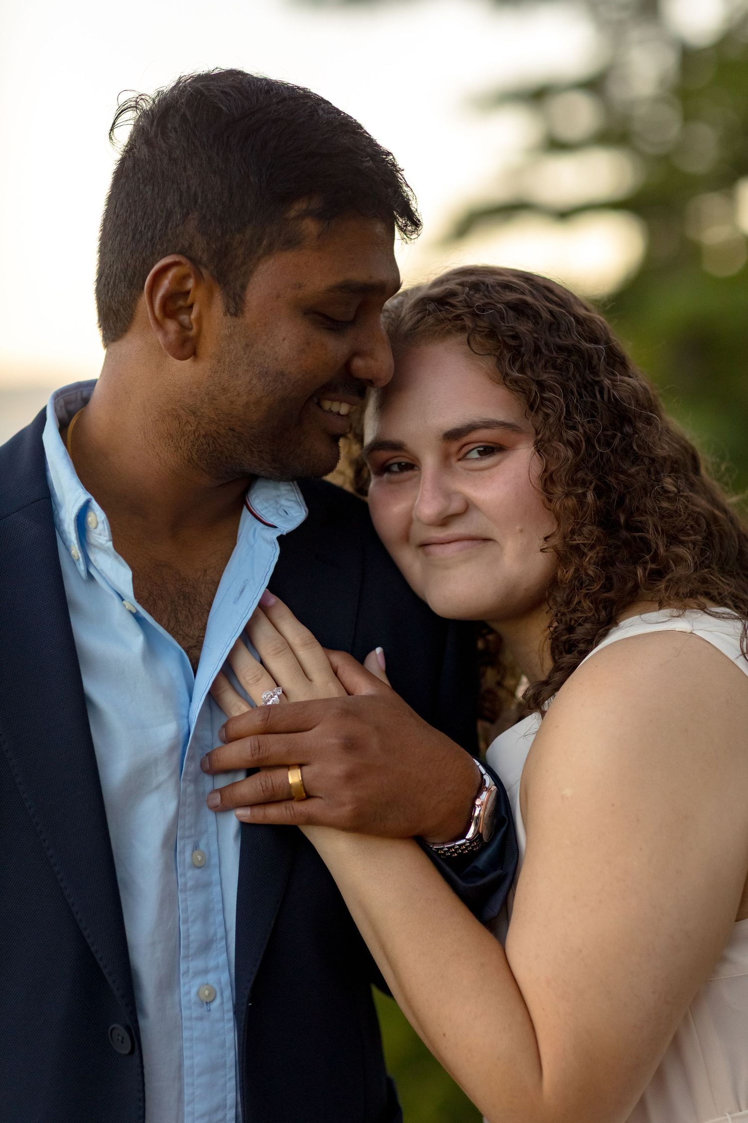 Romantic couple embracing outdoors at sunset, woman with curly hair smiling, wearing engagement ring.