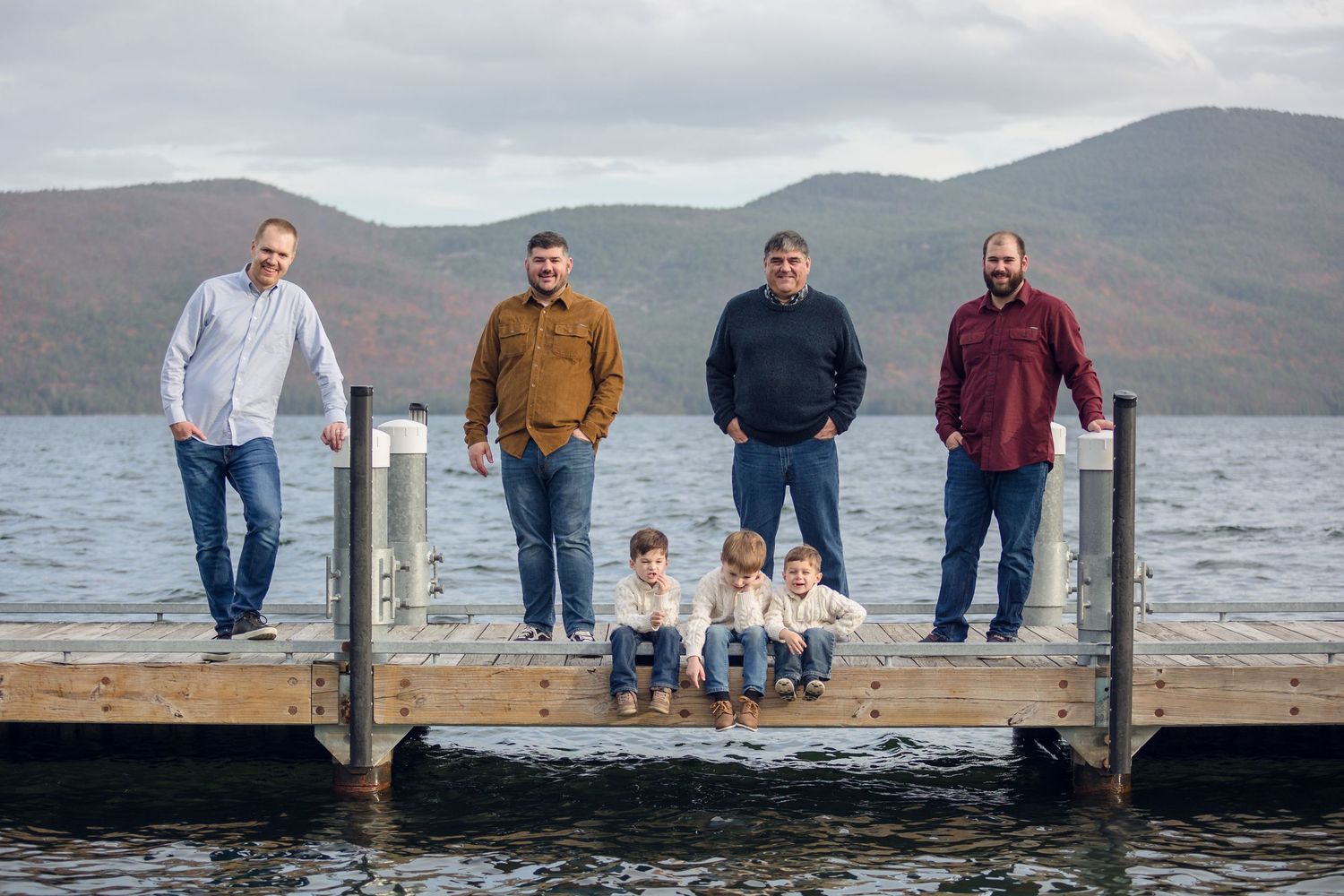 Group of people standing on a wooden pier overlooking a scenic mountain lake on a cloudy day.