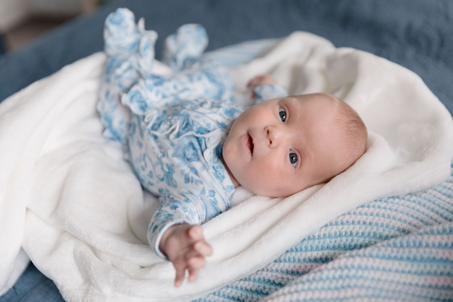 Baby in floral outfit lying on white bedding looking at camera.