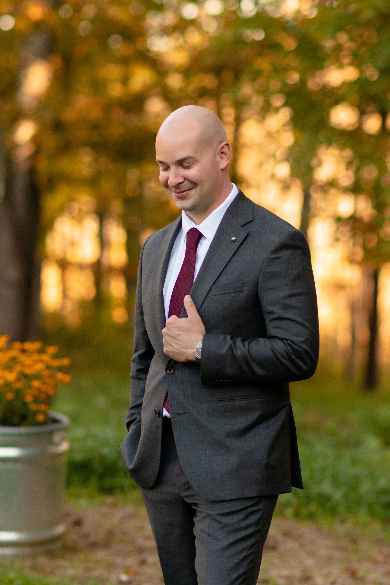Groom in dark suit and burgundy tie poses in autumn setting with golden sunlight.