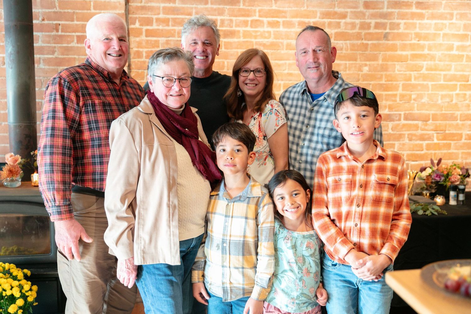 Multi-generational family group poses together against brick wall with yellow flowers in foreground.