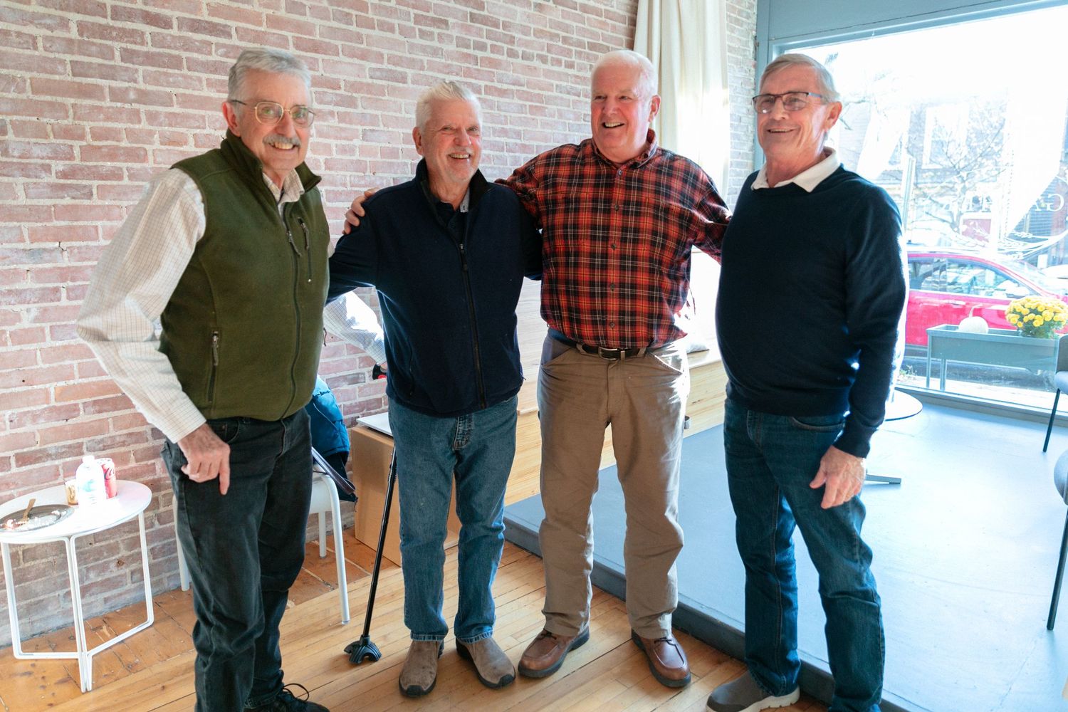 Four men in casual attire stand together against brick wall in social gathering setting.